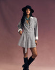 A person in the Xirena Louie Dress, a striped cotton mini shirtdress with removable self-tie belt, layered necklaces, black hat, and black knee-high boots stands against a plain mauve background.