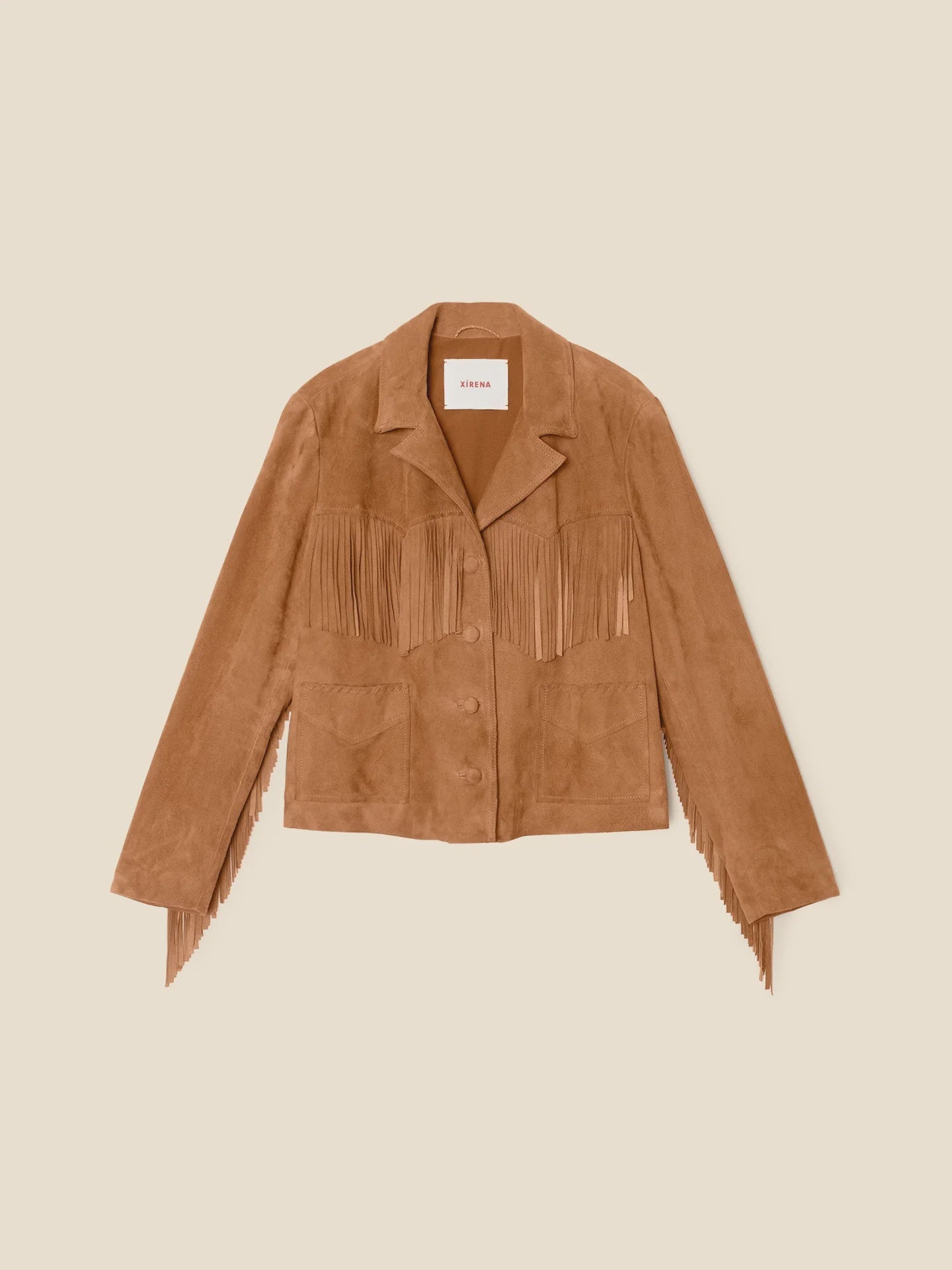 The Bronco Jacket by Xirena is a tan western fringe jacket with chest, sleeve, and pocket detailing. Made from goat suede, it features a notched collar and button-up front, displayed on a light beige background for a statement look.