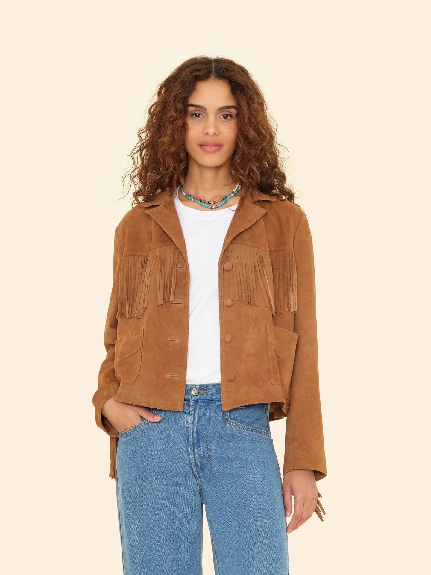 A woman with curly hair wears the Xirena Bronco Jacket in tan suede over a white tee and blue jeans, standing against a plain beige background.