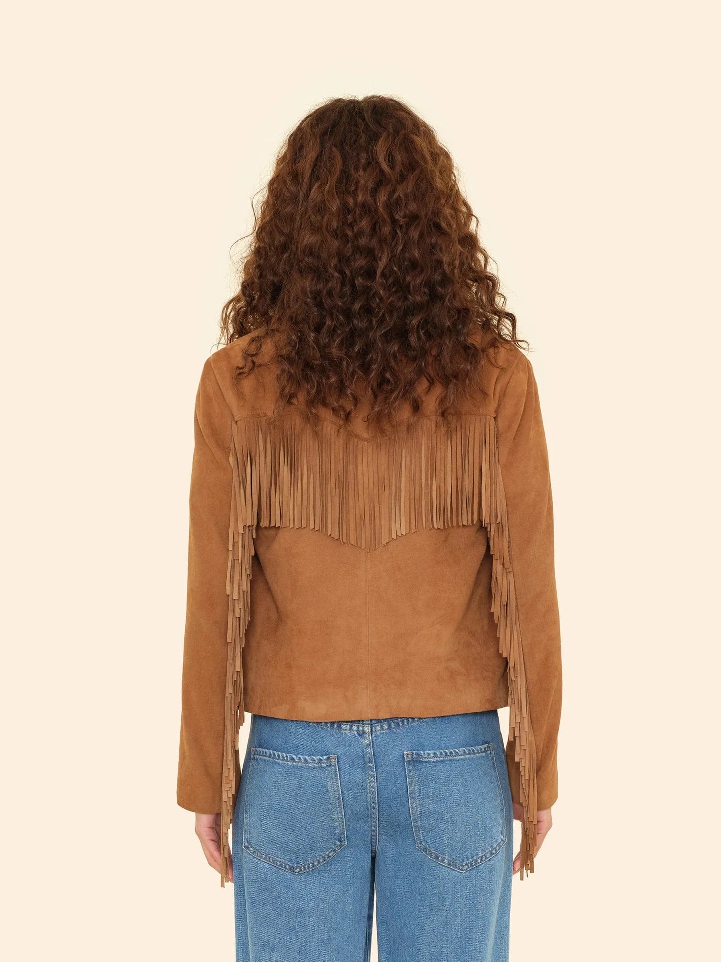 Seen from behind, a person with curly brown hair wears the Xirena Bronco Jacket in tan suede with long fringe and blue jeans, standing against a plain beige background.