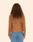 Seen from behind, a person with curly brown hair wears the Xirena Bronco Jacket in tan suede with long fringe and blue jeans, standing against a plain beige background.
