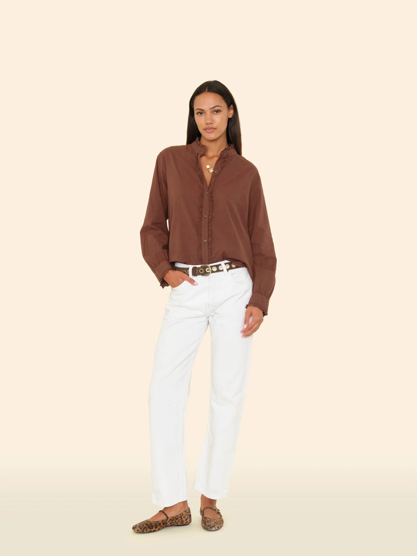 A woman poses against a beige background wearing the Xirena Amelie Shirt—a brown long sleeve button-up with subtle ruffle details—paired with white pants, a patterned belt, and leopard print flats, one hand in her pocket.