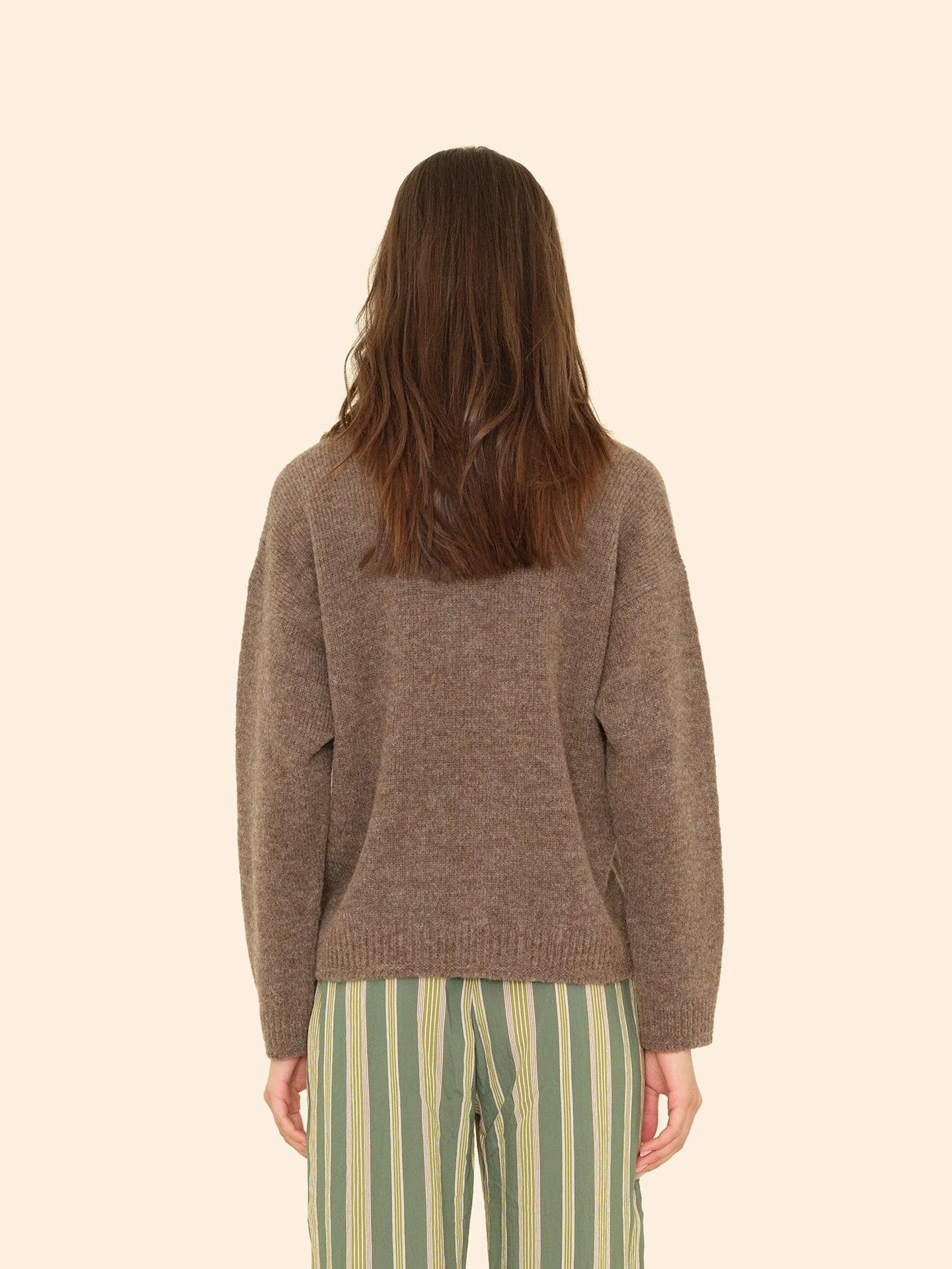 A person with long brown hair, seen from behind, wears the Xirena Ethan Sweater and green pants with vertical white and yellow stripes, standing against a beige background.