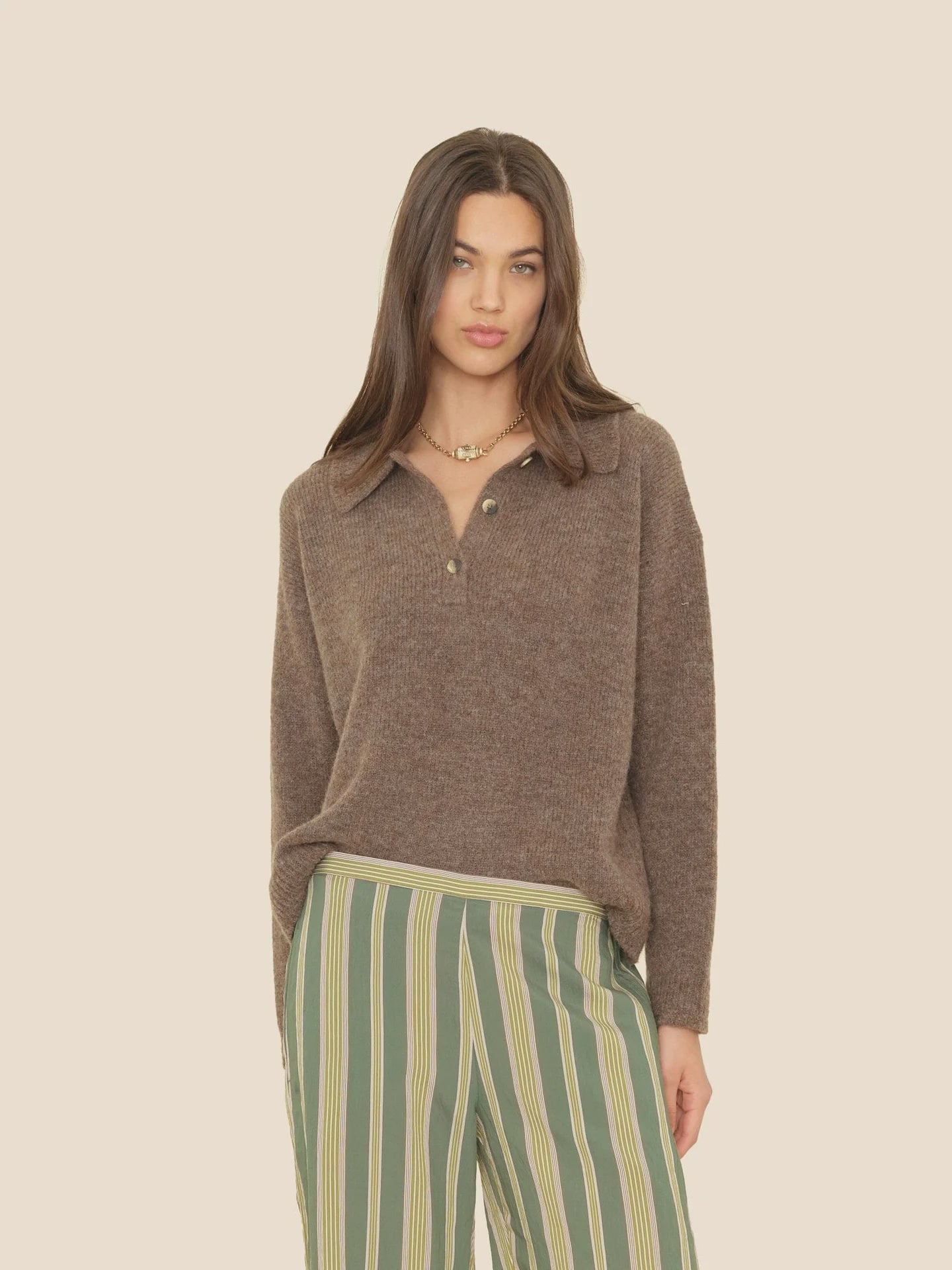 A woman wears the brown Ethan Sweater by Xirena, featuring buttons, paired with green pants with vertical cream and yellow stripes, standing against a plain beige background.