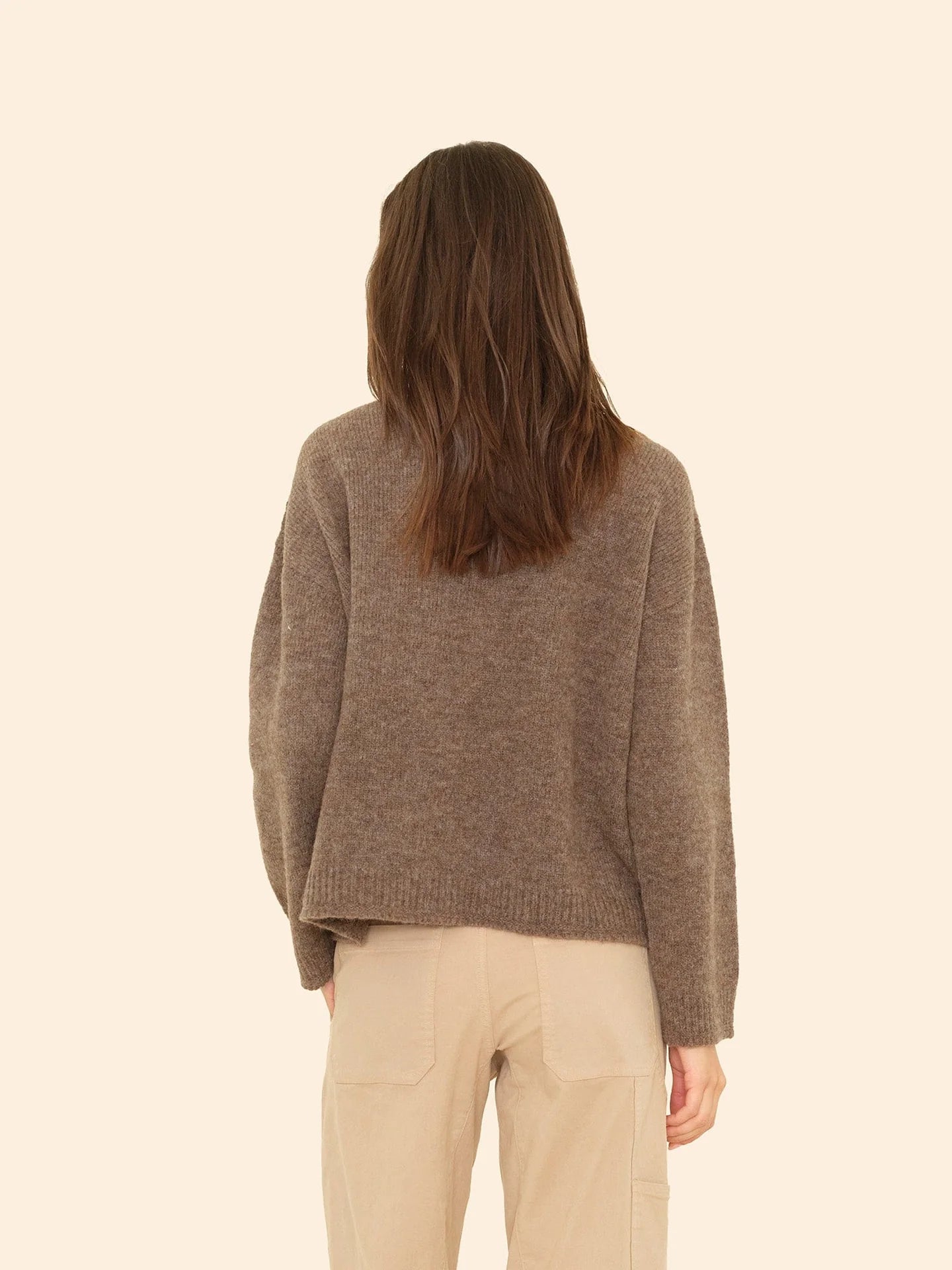 A woman with brown, shoulder-length hair is seen from the back, wearing the Xirena Julien Sweater and beige pants against a plain beige background.