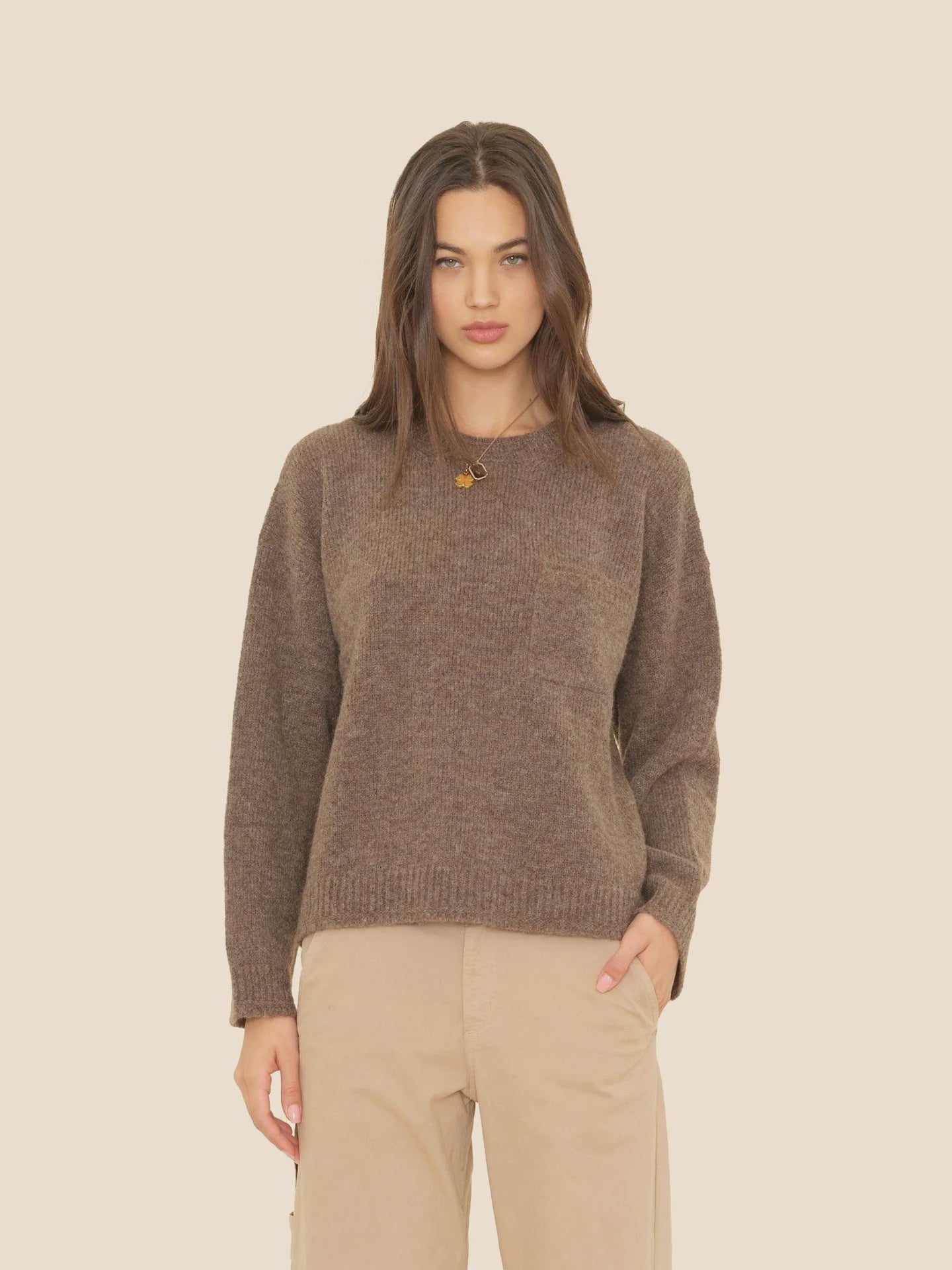 A woman with long brown hair models the Xirena Julien Sweater—a loose merino wool pullover—paired with beige pants, standing against a plain light background with one hand in her pocket.
