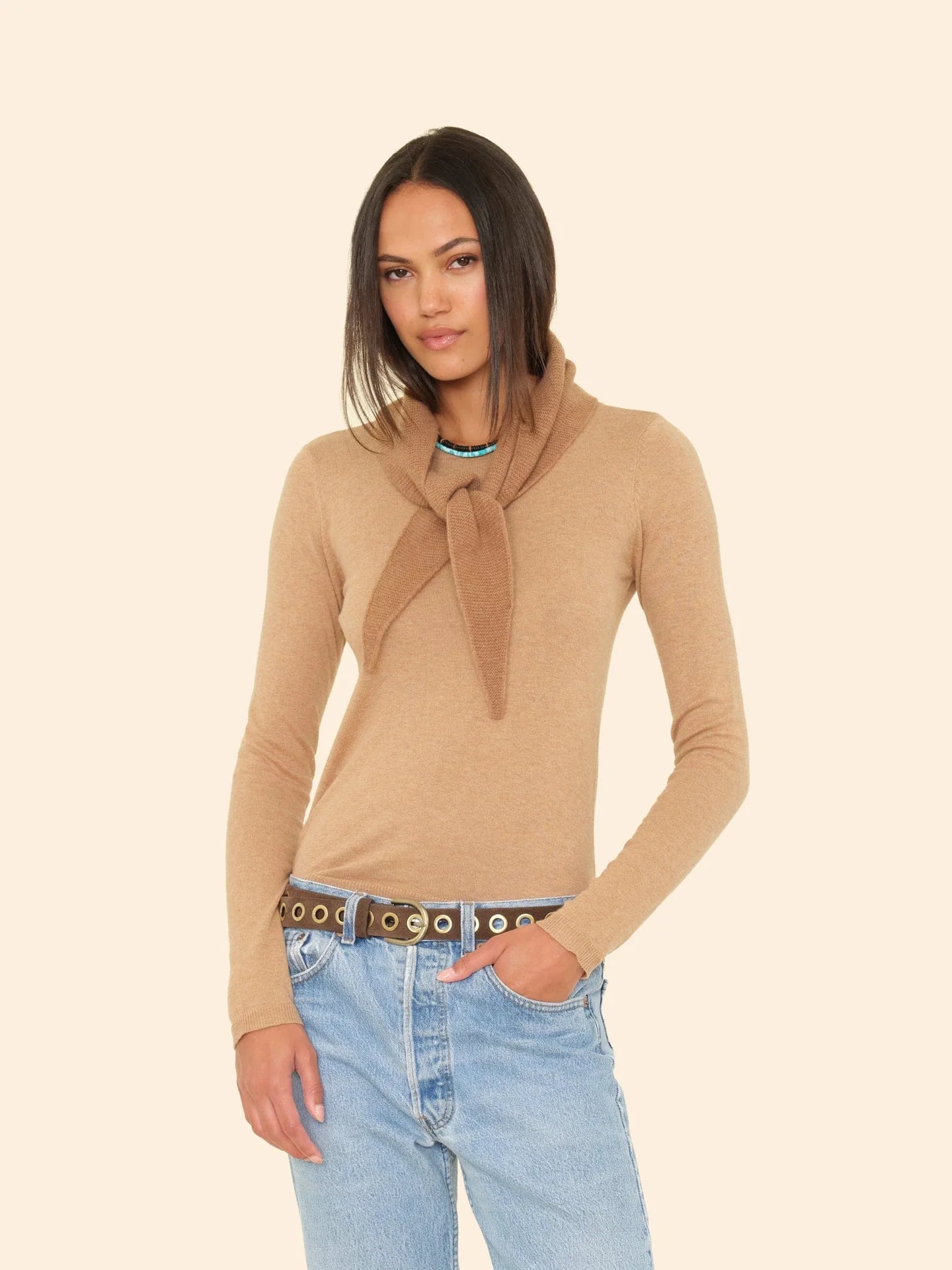 A woman with straight, dark hair stands against a beige background, wearing a tan sweater and light blue jeans, accessorized with the ultra-soft Myla Scarf by Xirena draped over her shoulders and a black belt with silver eyelets.