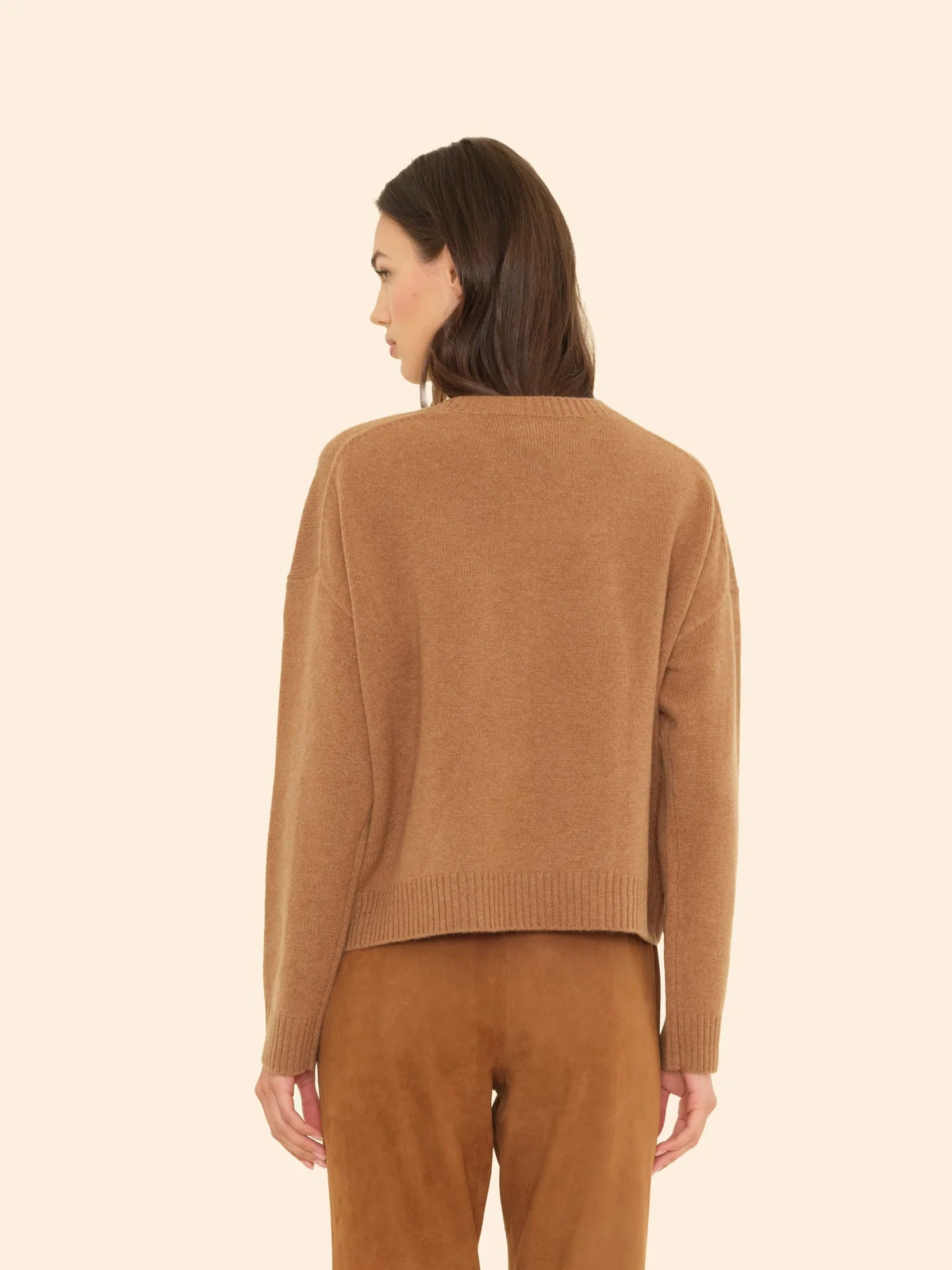A woman with shoulder-length brown hair faces away, wearing the ultra-soft Xirena Lela Sweater and matching suede pants against a light beige background.