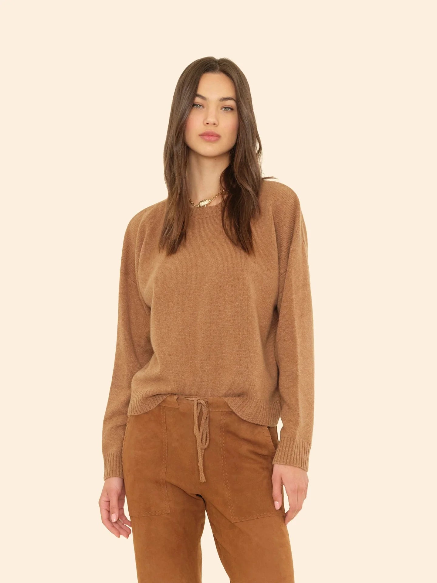 A woman with long brown hair models the Xirena Lela Sweater and matching tan pants, standing against a plain beige background.