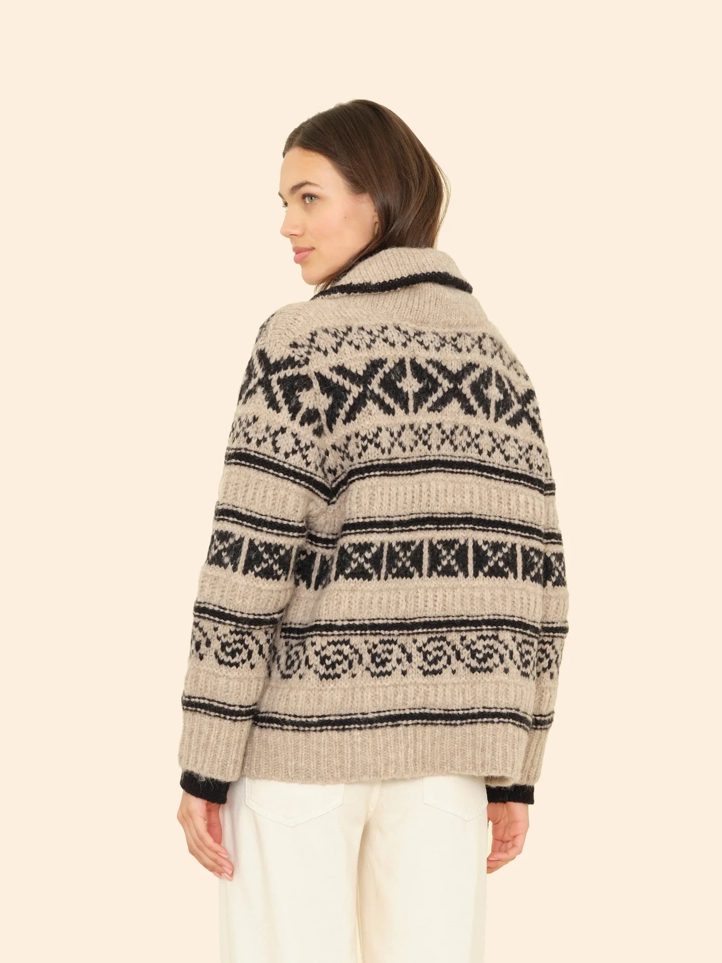 A woman with shoulder-length brown hair wears the Xirena Vail Cardigan—a beige and black alpaca sweater with a shawl collar—paired with white pants, standing against a plain light background and looking over her shoulder.