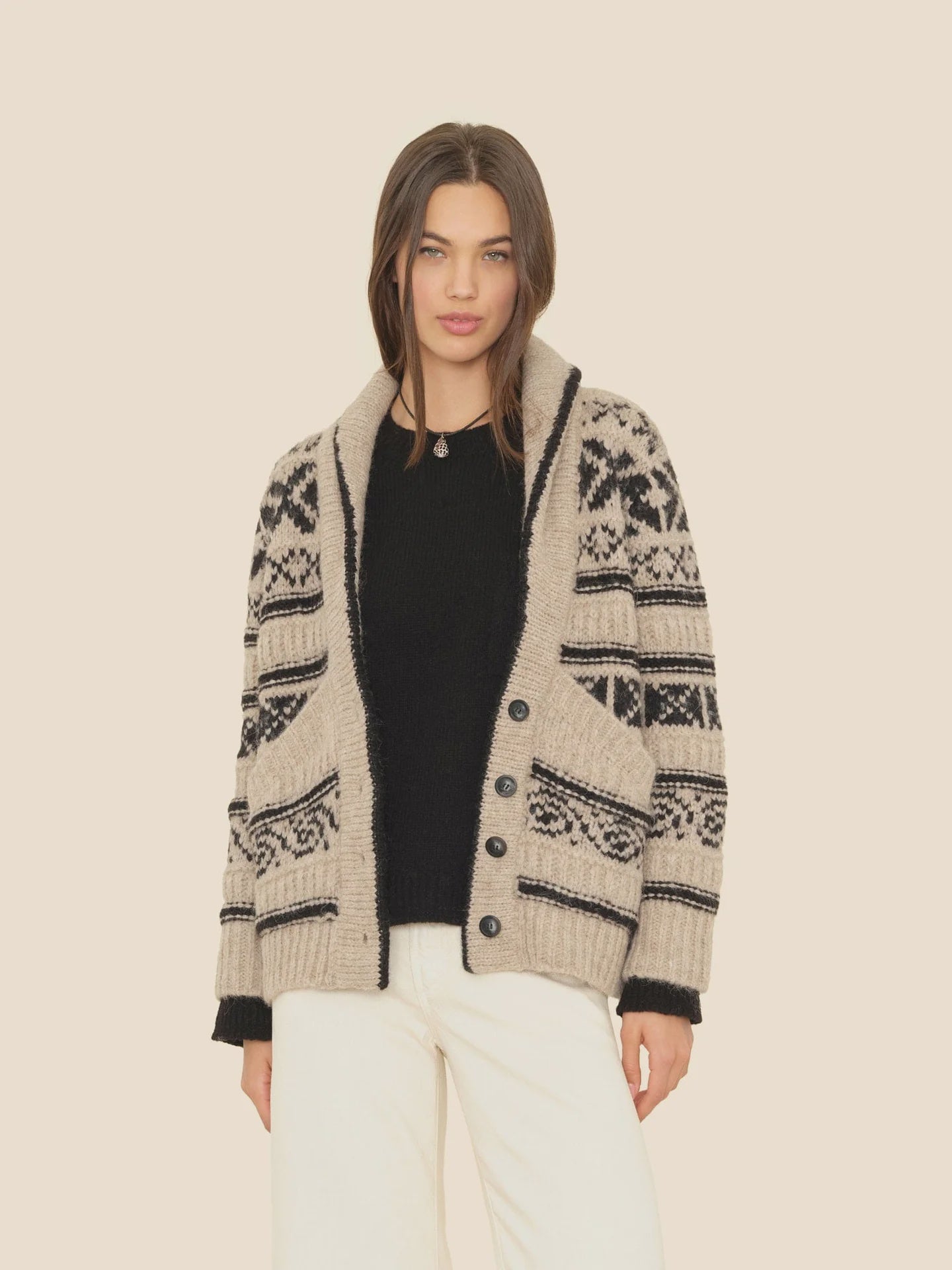 A woman wears the Xirena Vail Cardigan—beige with black geometric patterns—over a black sweater and white pants, standing against a plain light background.