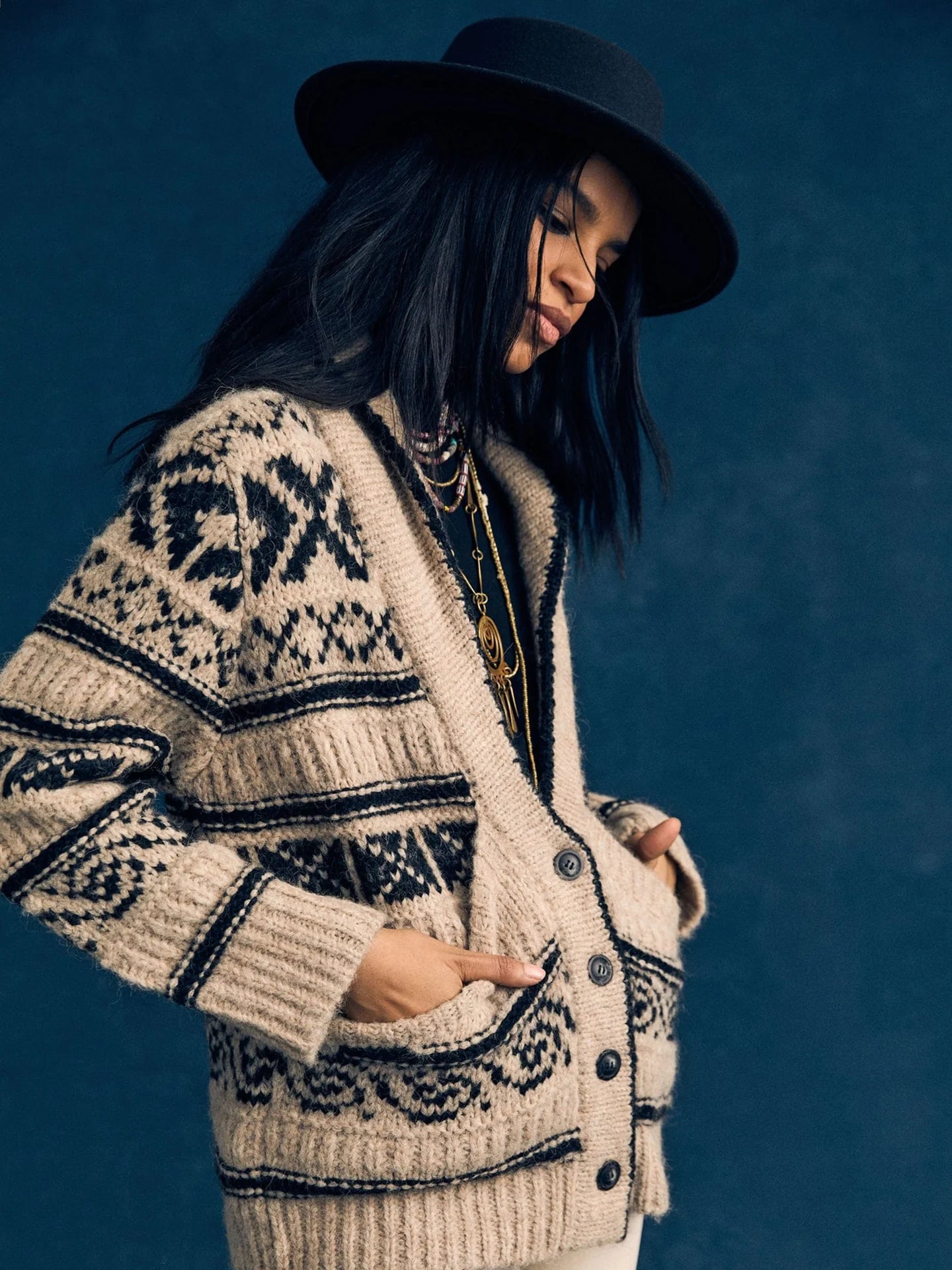 A woman wears the Xirena Vail Cardigan, layered necklaces, and a black wide-brim hat while standing against a dark blue background with her hands in her pockets, gazing downward in thought.