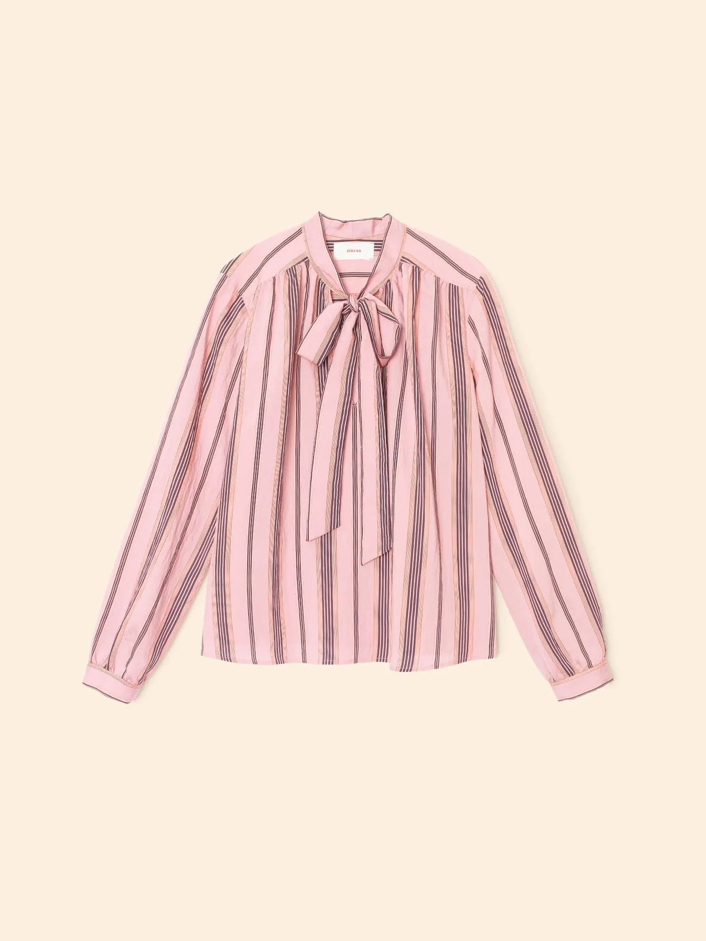 The Sabina Top by Xirena is a long-sleeved blouse with pink, black, and white vertical stripes, made from drapey viscose and finished with a chic bow tie neck, shown against a light beige background.