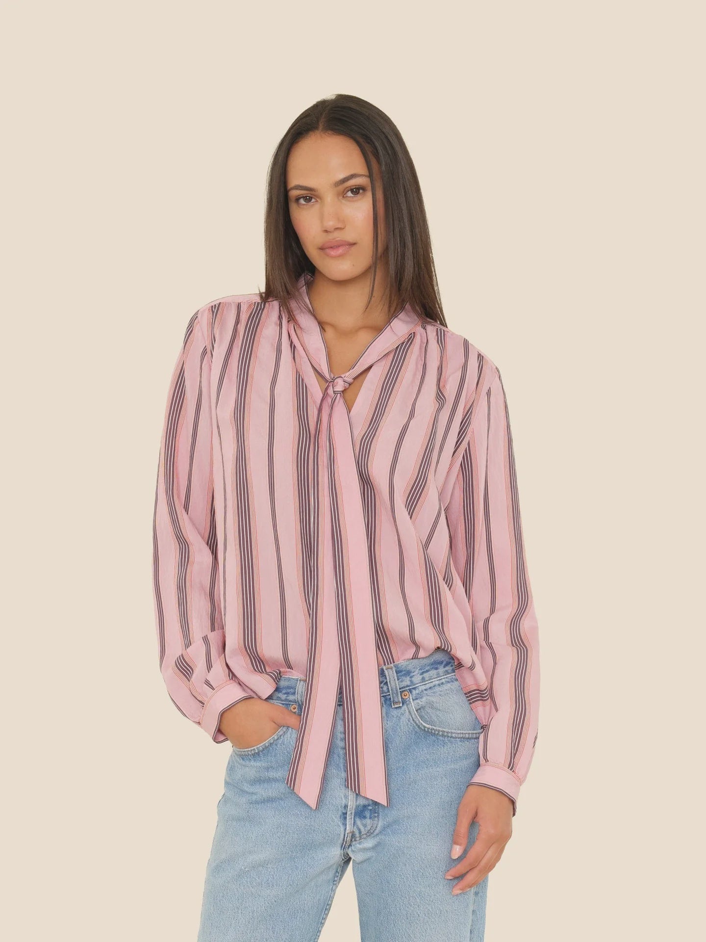 A woman wears the Sabina Top by Xirena—a drapey viscose blouse with black vertical stripes, a front tie, and deep V-neck—paired with light blue jeans, standing against a plain beige background.