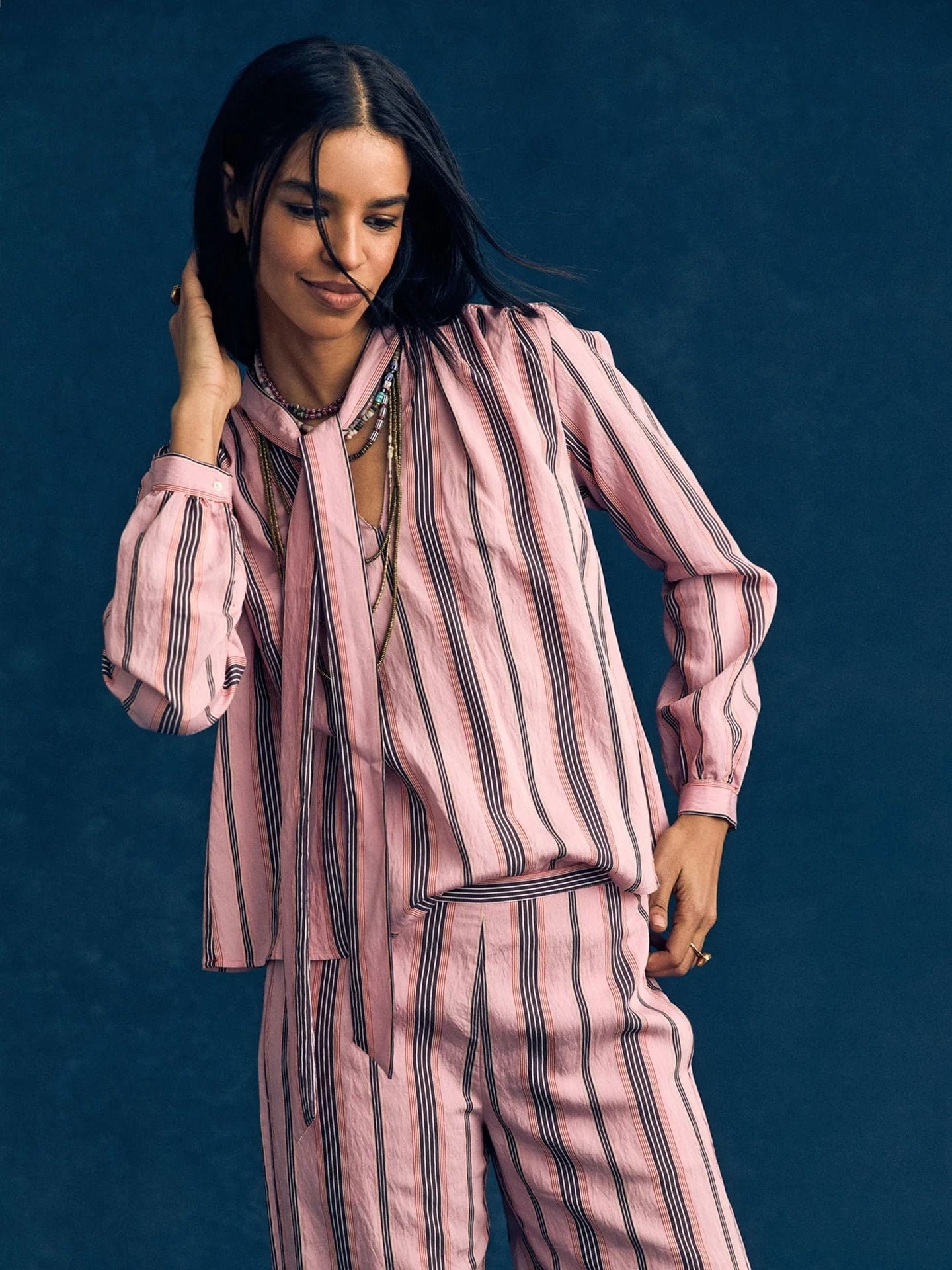 A woman with long dark hair smiles and touches her hair while wearing the Xirena Sabina Top—a drapey pink and black vertical striped blouse—paired with a matching scarf and pants, set against a dark blue background.
