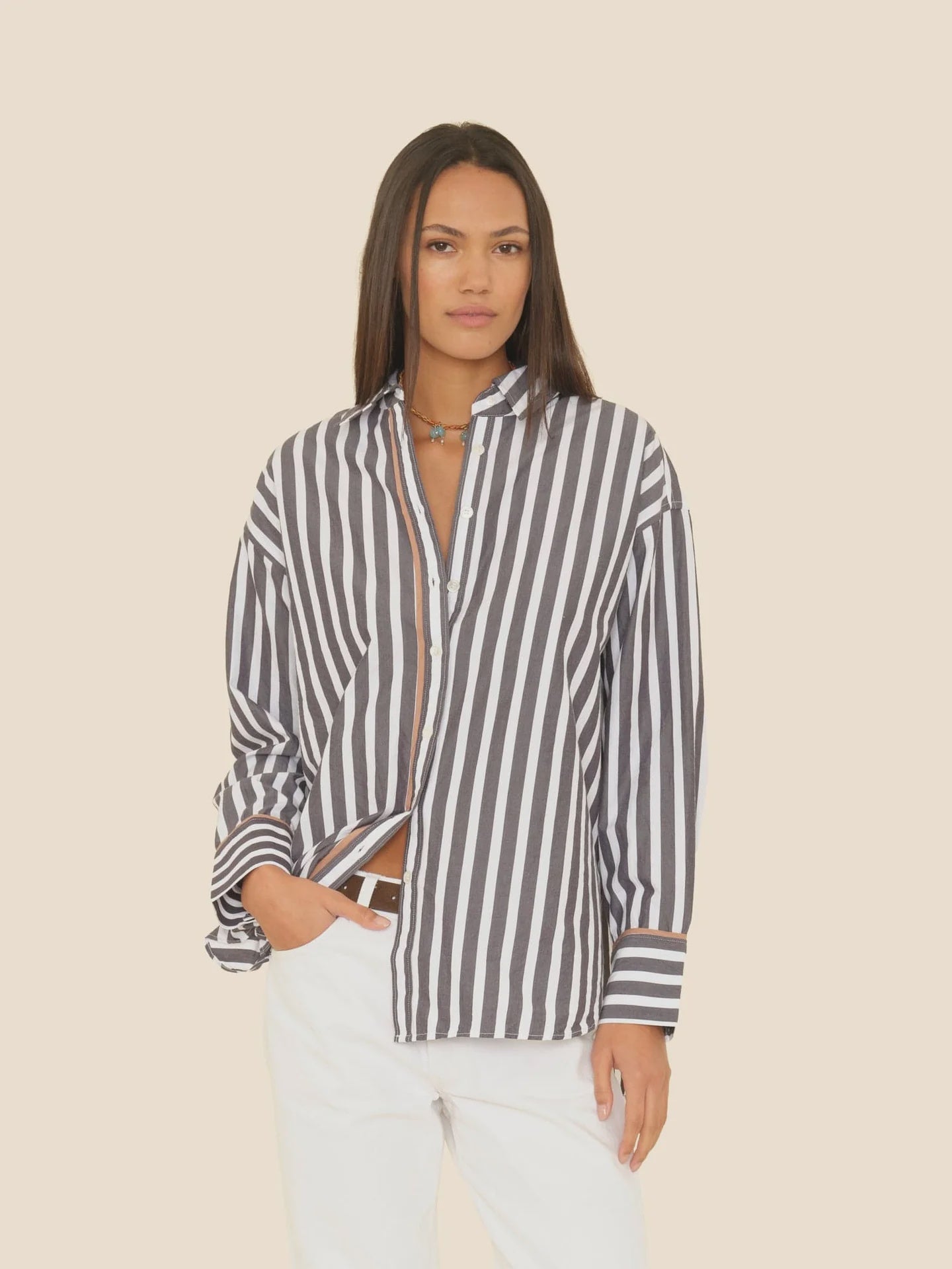A woman with long straight hair and a neutral expression wears the Xirena Gillian Shirt, a relaxed black-and-white striped cotton button-front, paired with white pants against a plain beige background.