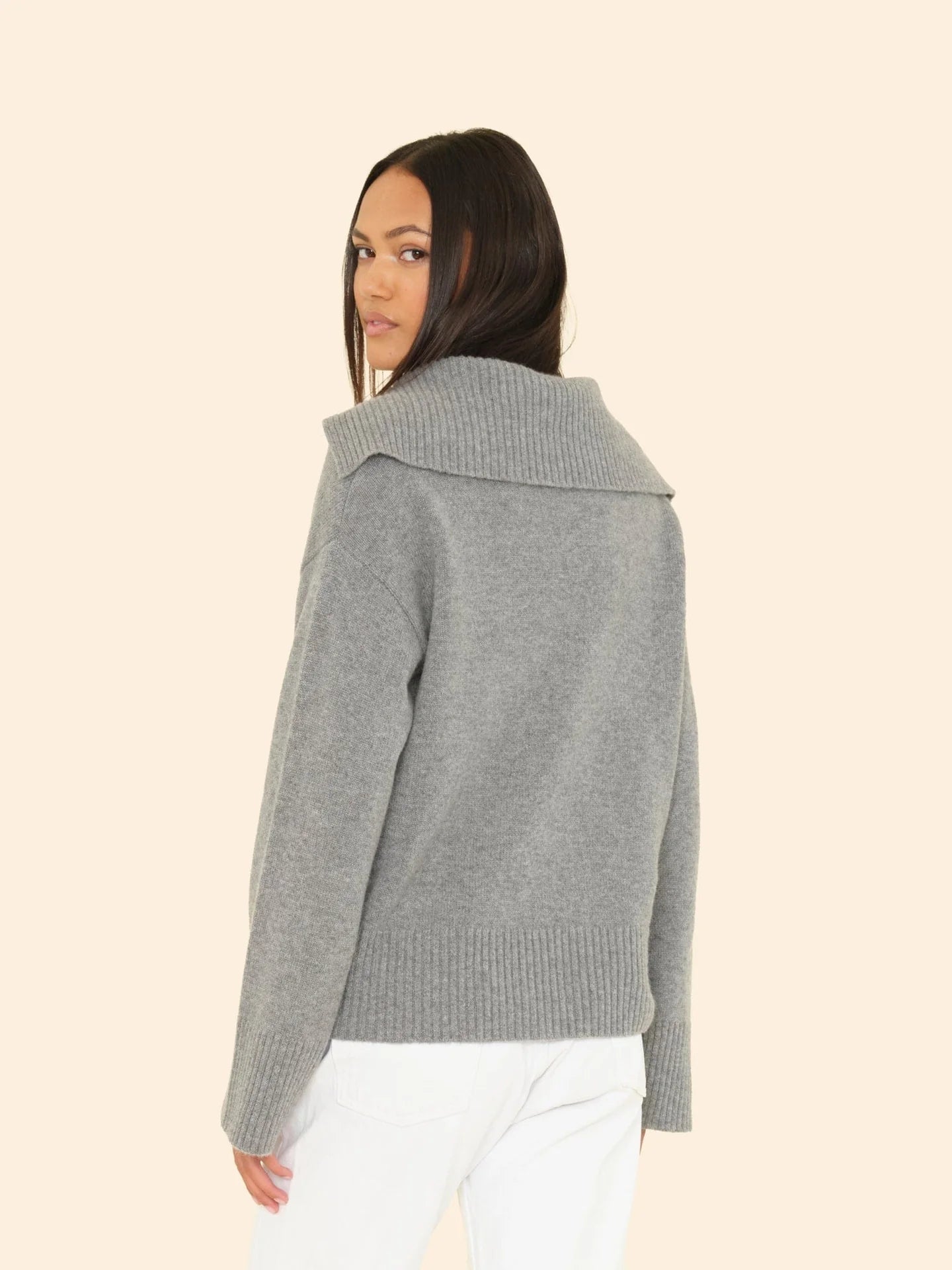 A woman with long dark hair wears the Xirena Mattia Sweater, a luxe cashmere pullover with a wide collar, paired with white pants. She stands against a light beige background and glances over her shoulder at the camera.