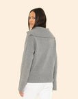 A woman with long dark hair wears the Xirena Mattia Sweater, a luxe cashmere pullover with a wide collar, paired with white pants. She stands against a light beige background and glances over her shoulder at the camera.