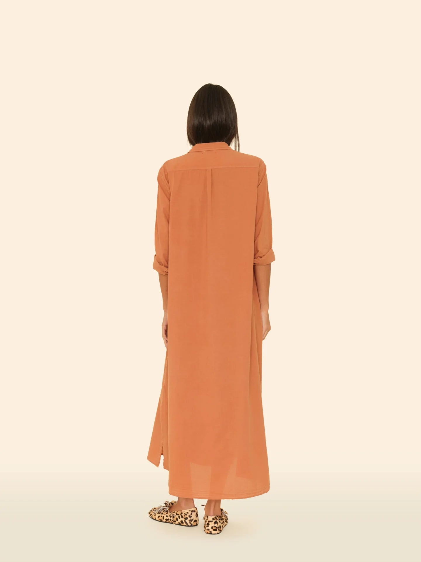A person with straight dark hair faces away, wearing the Xirena Boden Dress—an orange cotton poplin shirtdress with rolled-up sleeves—paired with leopard print slip-on shoes, set against a light beige background.