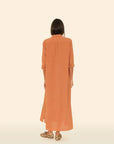 A person with straight dark hair faces away, wearing the Xirena Boden Dress—an orange cotton poplin shirtdress with rolled-up sleeves—paired with leopard print slip-on shoes, set against a light beige background.