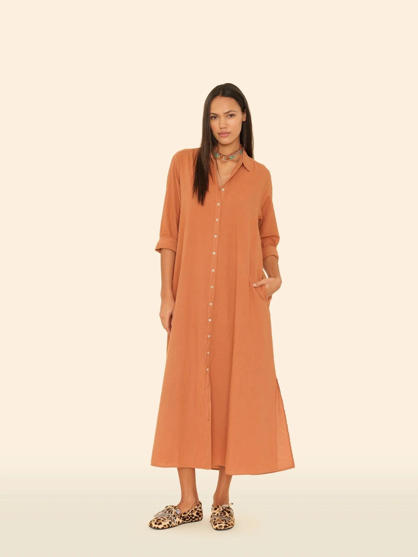 A woman stands against a beige background wearing the Xirena Boden Dress, a long, loose cotton poplin shirtdress in rust with rolled-up sleeves and her hand in the pocket. She pairs it with leopard print slip-on shoes and has long straight hair.