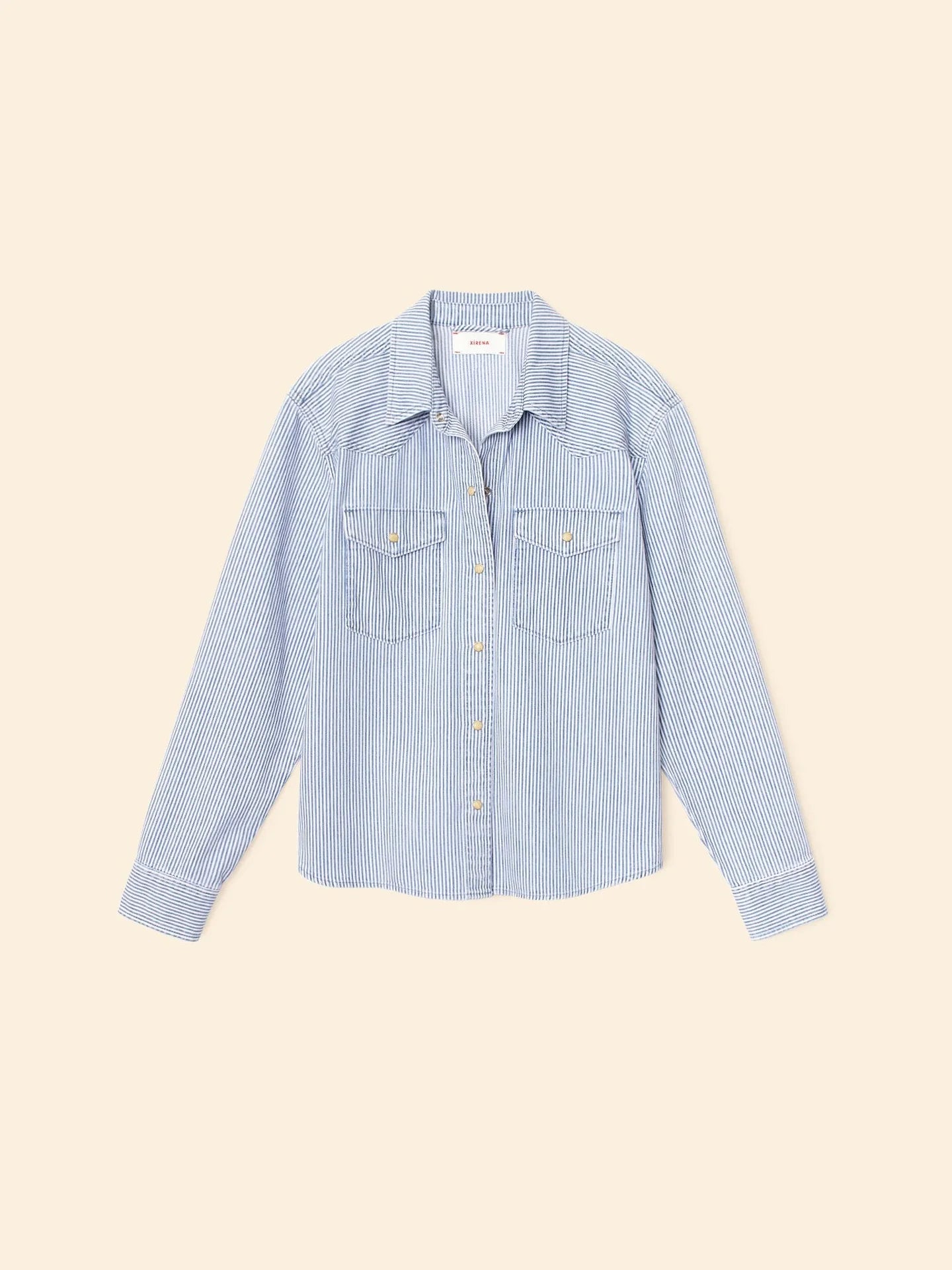 The Byron Shirt by Xirena is a light blue and white railroad striped denim button-front shirt with long sleeves, a collar, and two chest pockets, shown laid flat on a beige background.