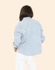A person with shoulder-length brown hair, seen from behind, wears the Xirena Byron Shirt—a blue and white railroad striped denim button-front top—with white pants against a plain beige background.