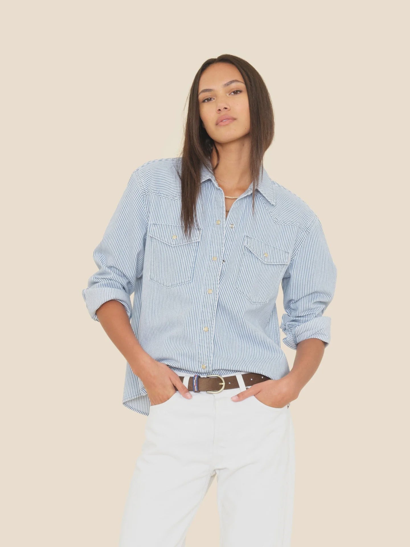 A person with long straight hair models the Xirena Byron Shirt in light blue with pearl snap buttons and white pants, posing with one hand in their pocket against a light beige background.