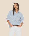 A person with long straight hair models the Xirena Byron Shirt in light blue with pearl snap buttons and white pants, posing with one hand in their pocket against a light beige background.