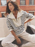 A woman with long brown hair sits outdoors in sunlight, wearing the Kat Sweatshirt by Xirena—a cream and navy striped cotton zip-collar top—paired with light pants. A brick building can be seen in the background.