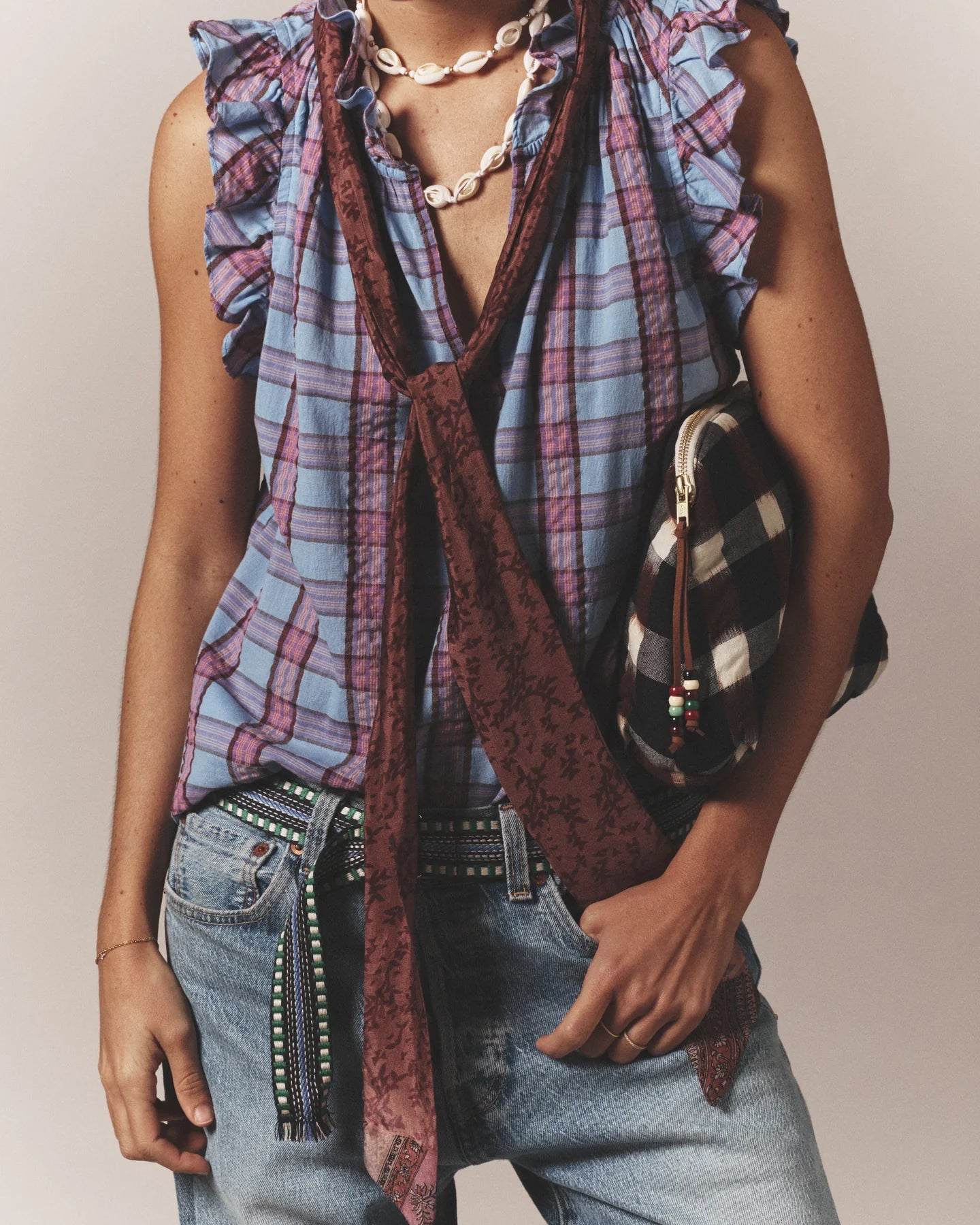 Person wearing a sleeveless, ruffled blue and pink plaid top, maroon scarf, Xirena Stevie Belt, light blue jeans, shell necklace, and carrying a black-and-white checkered bag.