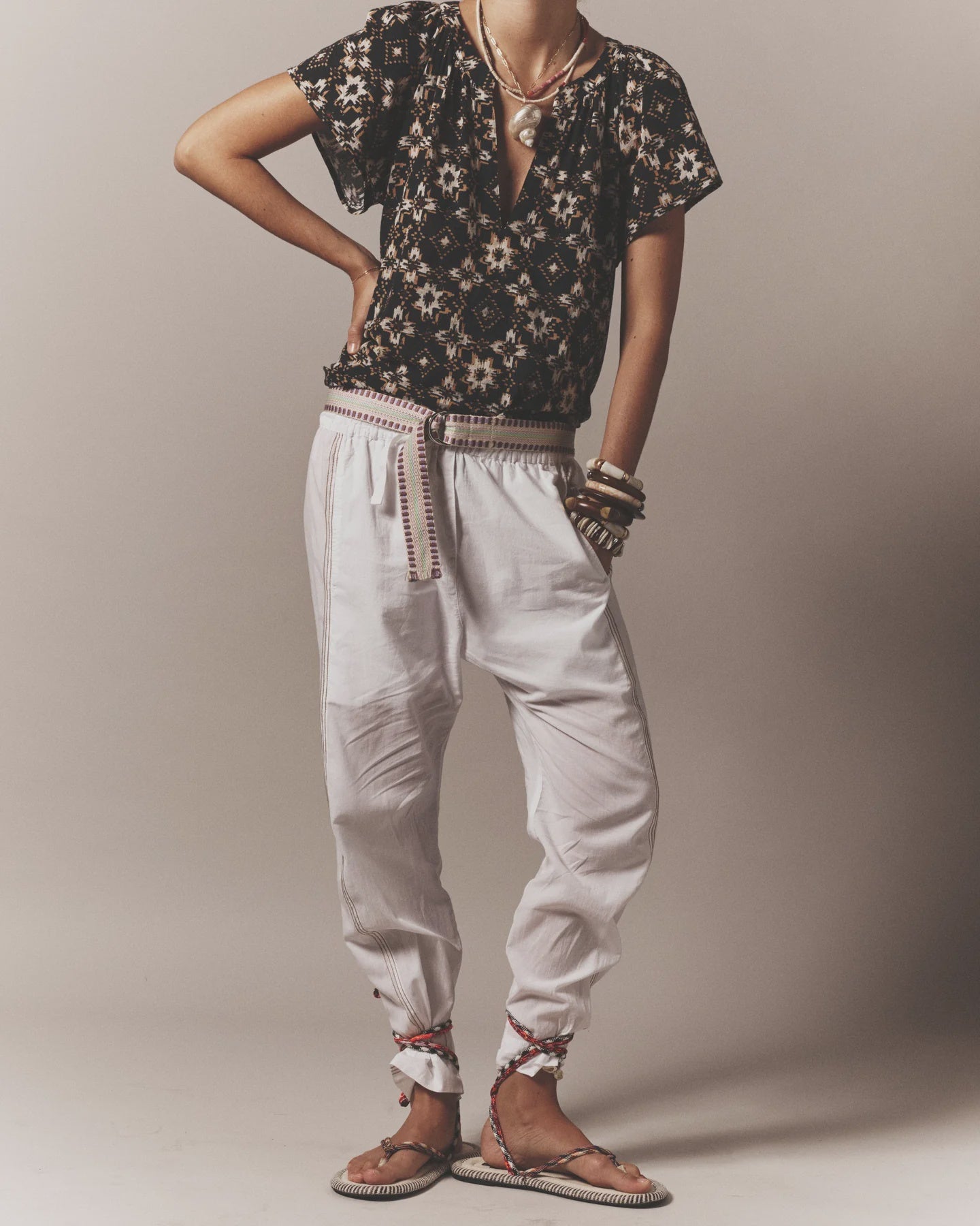 A person poses in a black patterned blouse, loose white pants with the Stevie Belt by Xirena, ankle-tied espadrille sandals, and several bracelets against a neutral background. Their head is not shown in the image.