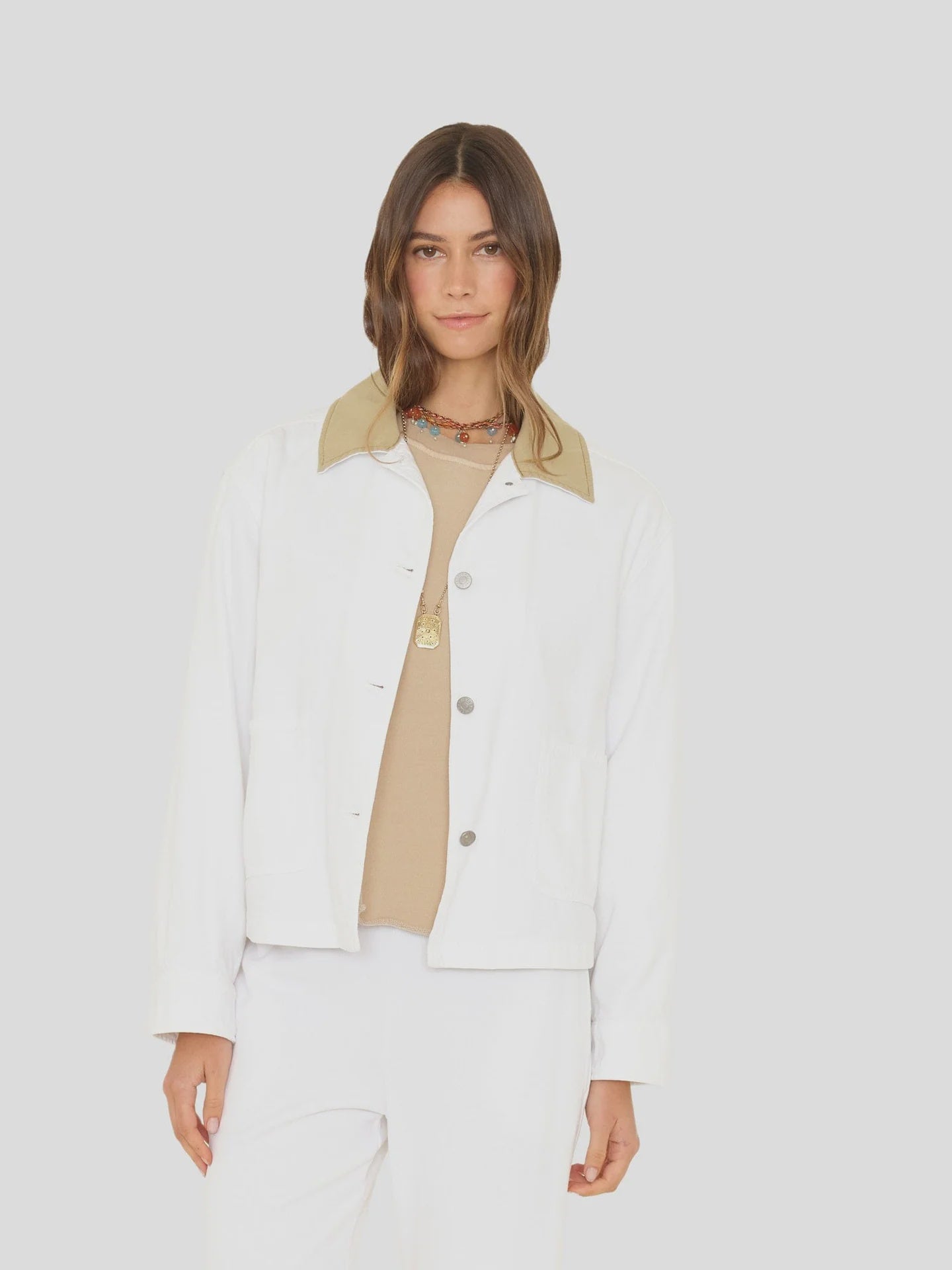 A woman with long brown hair wears the Xirena Cove Jacket—a white lyocell denim piece with a beige collar—over a beige shirt and white pants, standing against a light gray background. The jacket is made in Los Angeles.