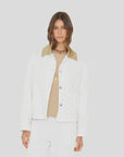 A woman with long brown hair wears the Xirena Cove Jacket—a white lyocell denim piece with a beige collar—over a beige shirt and white pants, standing against a light gray background. The jacket is made in Los Angeles.