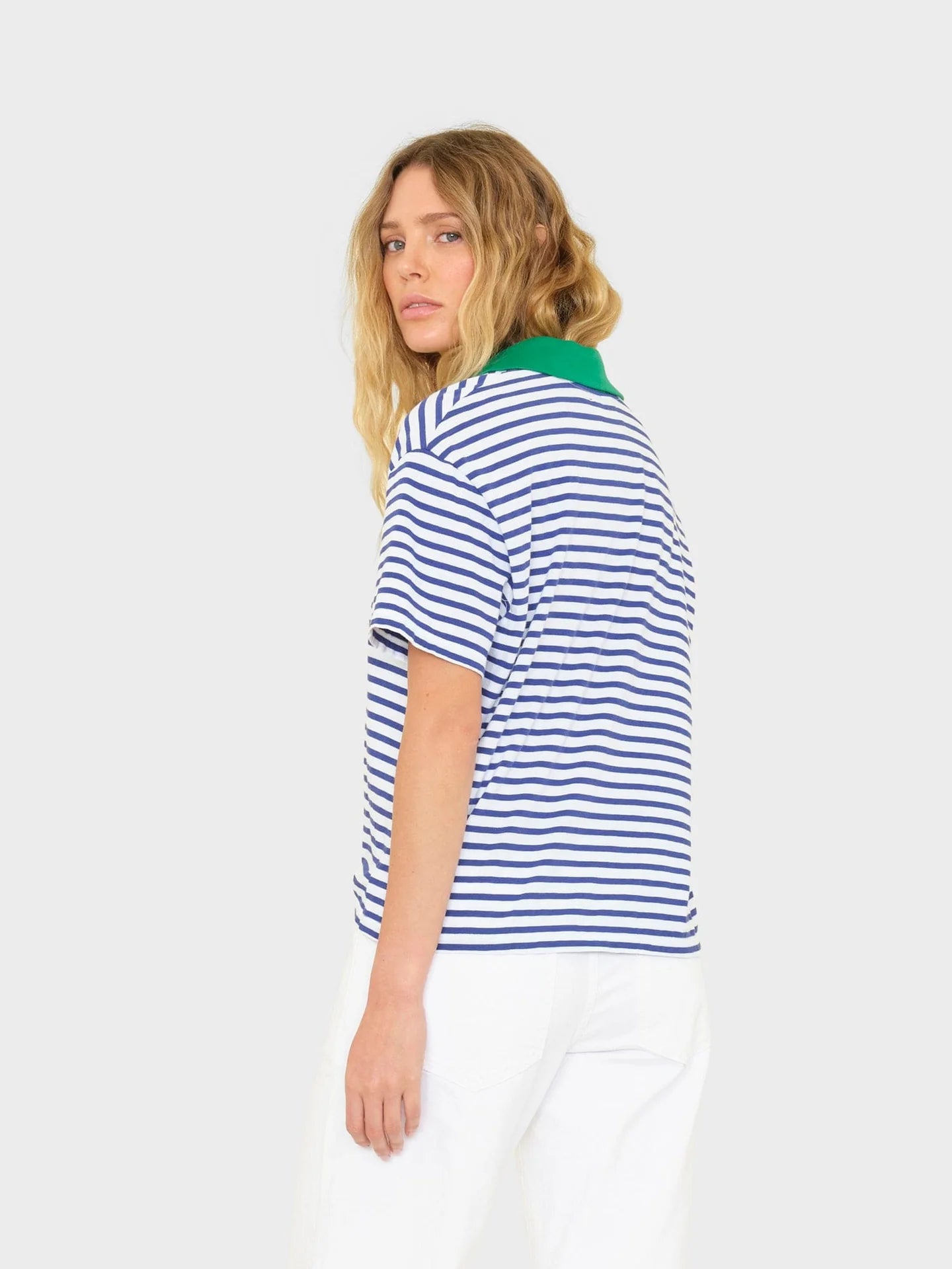 A person with wavy blonde hair stands against a plain light gray background, wearing the Xirena Kellie Top—an ultra-soft blue and white striped cotton jersey short sleeve polo tee with a green collar—and white pants, looking over their shoulder.
