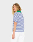 A person with wavy blonde hair stands against a plain light gray background, wearing the Xirena Kellie Top—an ultra-soft blue and white striped cotton jersey short sleeve polo tee with a green collar—and white pants, looking over their shoulder.