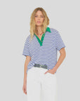 A woman with wavy blonde hair wears the Xirena Kellie Top, a blue and white striped cotton jersey polo tee with a green collar, tucked into white pants, standing against a plain light gray background.