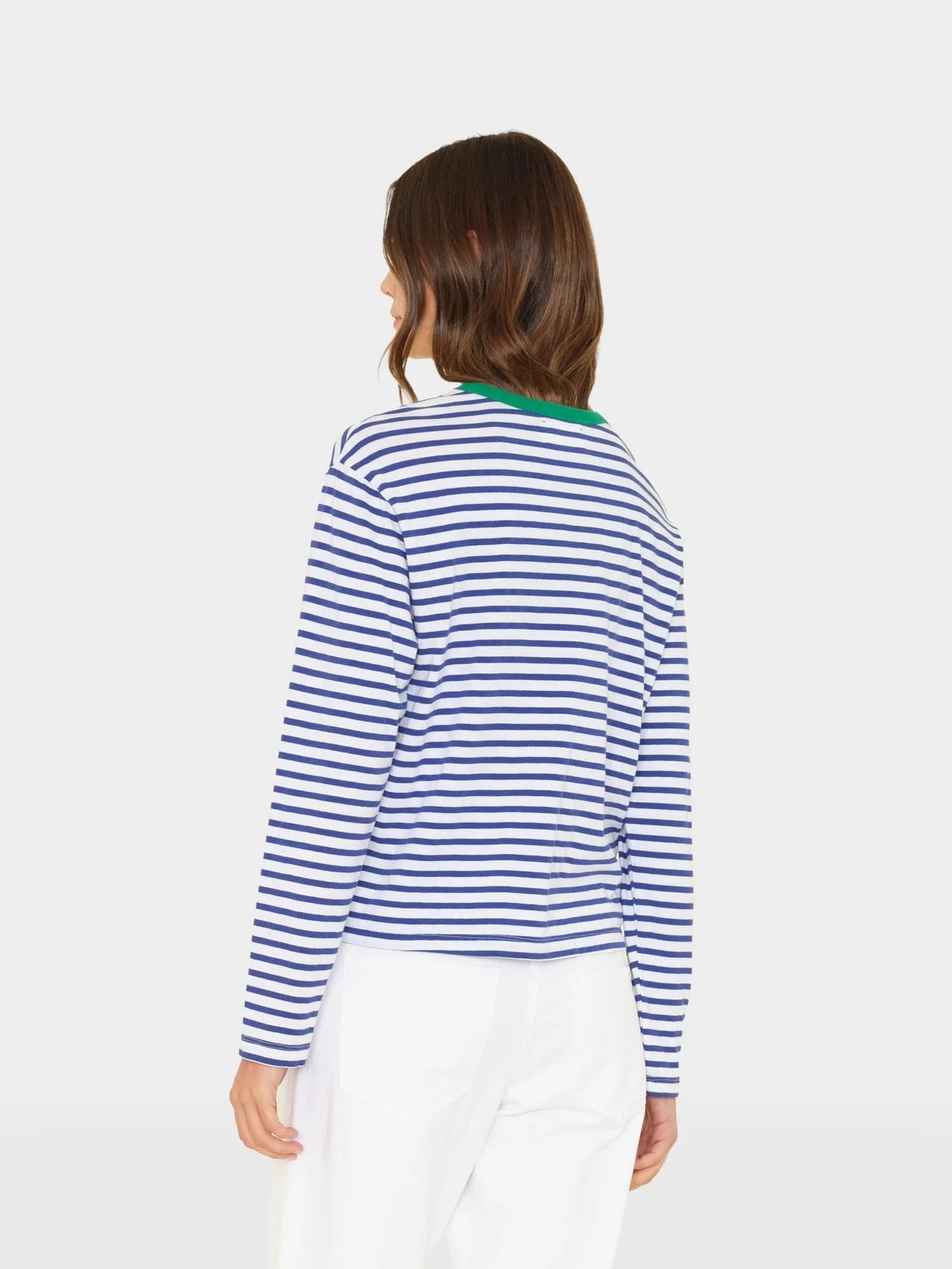 A person with shoulder-length brown hair is seen from the back wearing the Xirena Terrance Tee, a blue and white striped boxy-fit long sleeve cotton top with a green collar, paired with white pants against a plain light background.