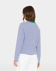 A person with shoulder-length brown hair is seen from the back wearing the Xirena Terrance Tee, a blue and white striped boxy-fit long sleeve cotton top with a green collar, paired with white pants against a plain light background.