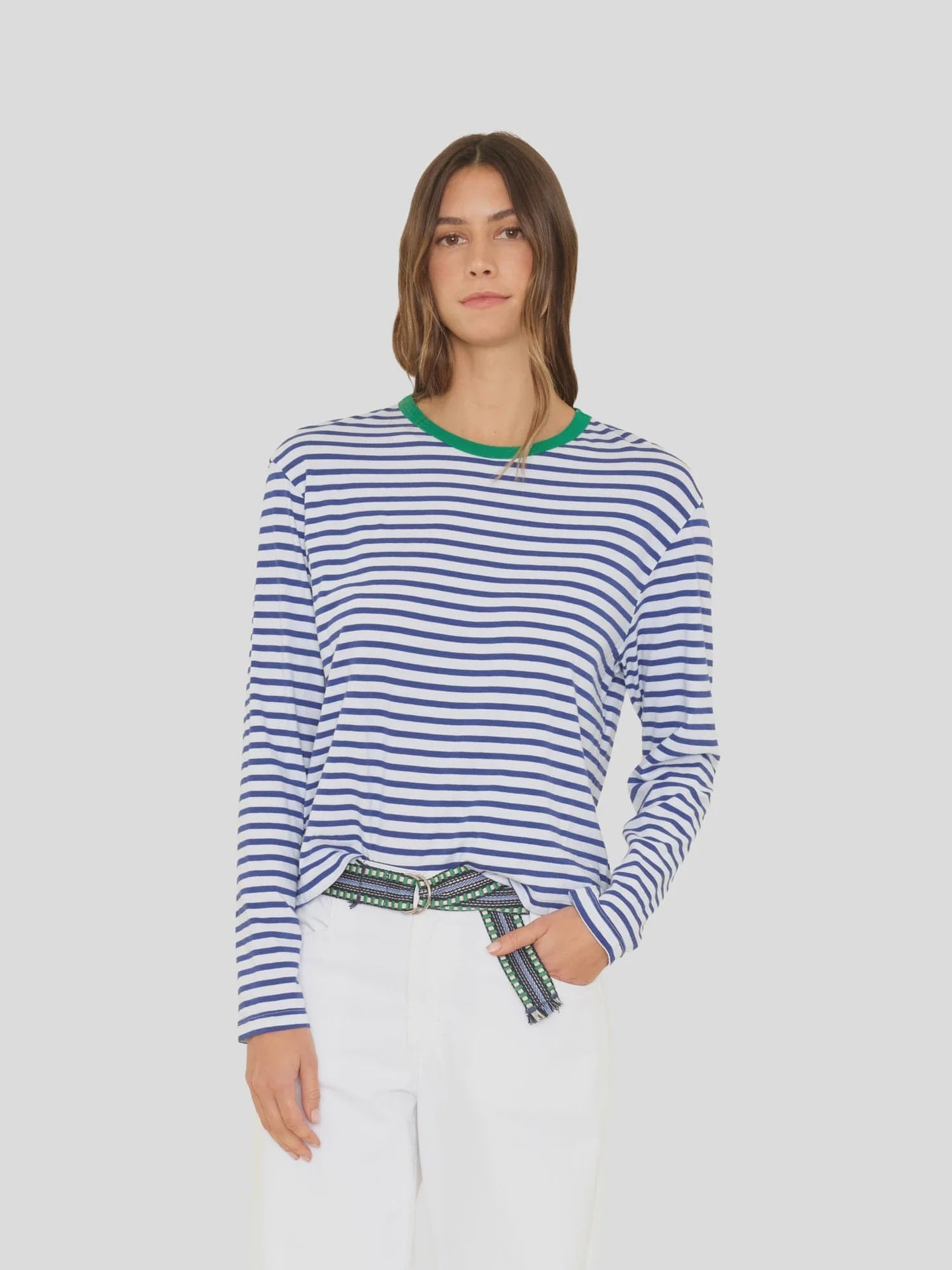 Wearing the Xirena Terrance Tee—a boxy fit cotton jersey long sleeve with blue and white stripes and a green neckline—a long-haired person pairs it with white pants and a black-green patterned belt against a plain light background.