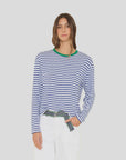 Wearing the Xirena Terrance Tee—a boxy fit cotton jersey long sleeve with blue and white stripes and a green neckline—a long-haired person pairs it with white pants and a black-green patterned belt against a plain light background.