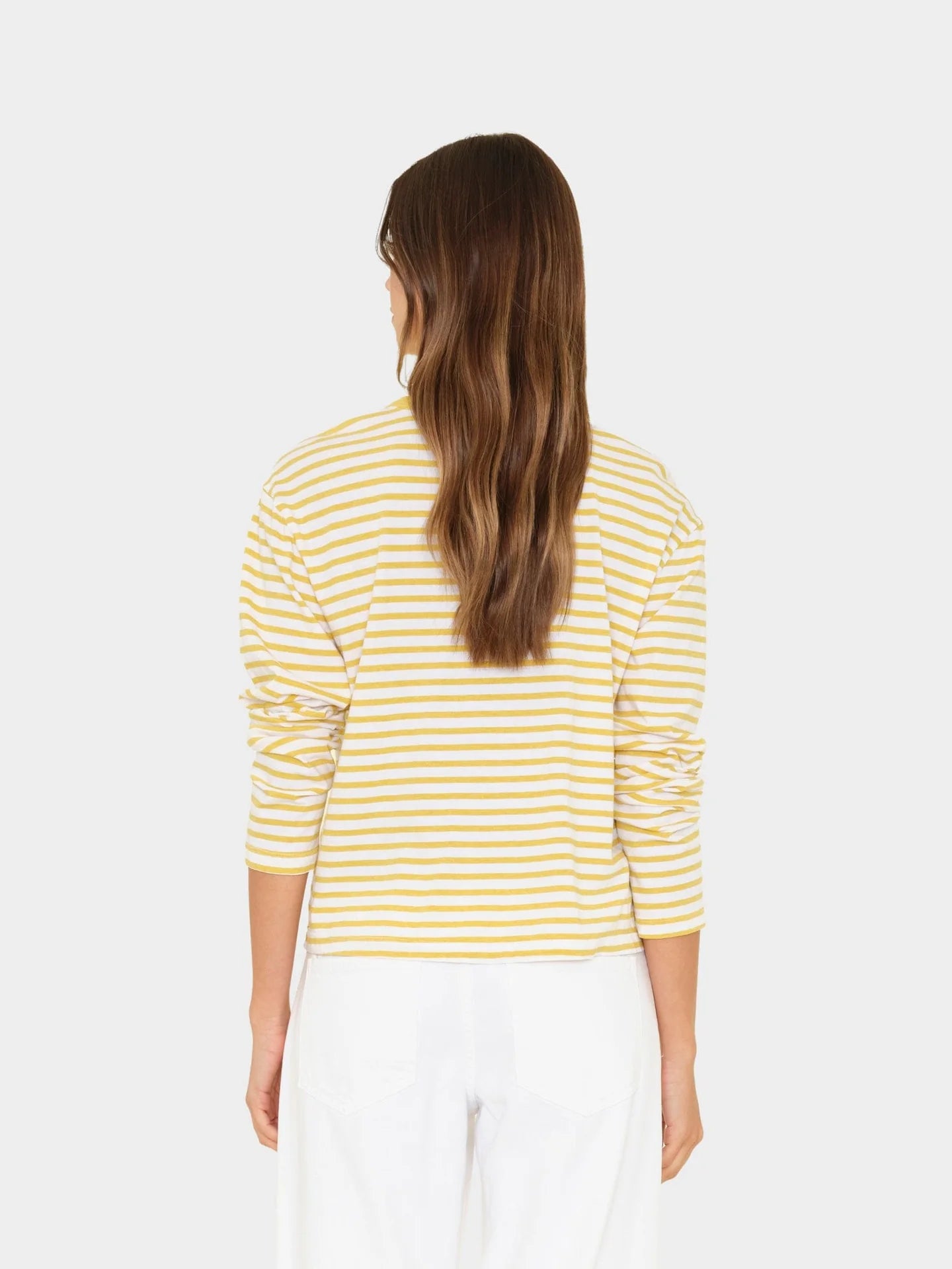 A person with long brown hair, seen from behind, wears the Xirena Terrance Top—a yellow and white striped cotton jersey long sleeve tee—and white pants against a plain light background.