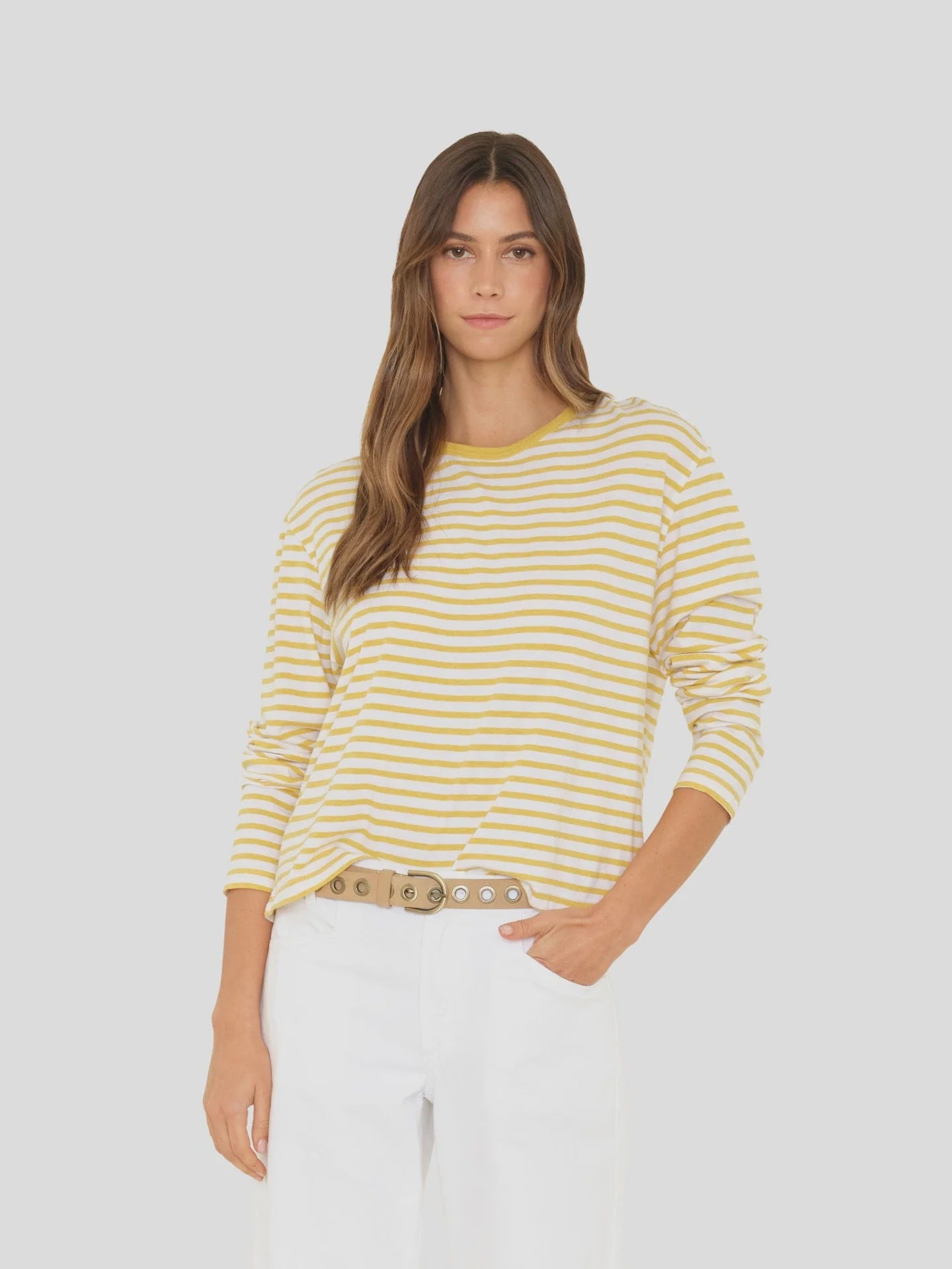 A woman with long brown hair wears the Xirena Terrance Top—a yellow and white striped cotton jersey long sleeve tee—paired with white pants and a beige belt, standing with one hand in her pocket against a plain light gray background.