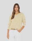 A woman with long brown hair wears the Xirena Terrance Top—a yellow and white striped cotton jersey long sleeve tee—paired with white pants and a beige belt, standing with one hand in her pocket against a plain light gray background.