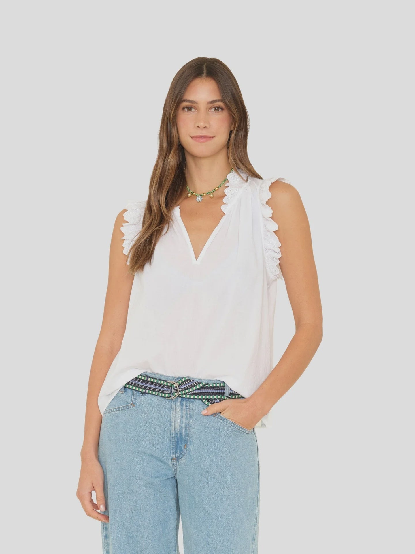 A woman wearing the Xirena Bexie Top, a white cotton gauze blouse with ruffle details, stands against a plain light gray background. She pairs it with light blue jeans, a beaded belt, and a green necklace.