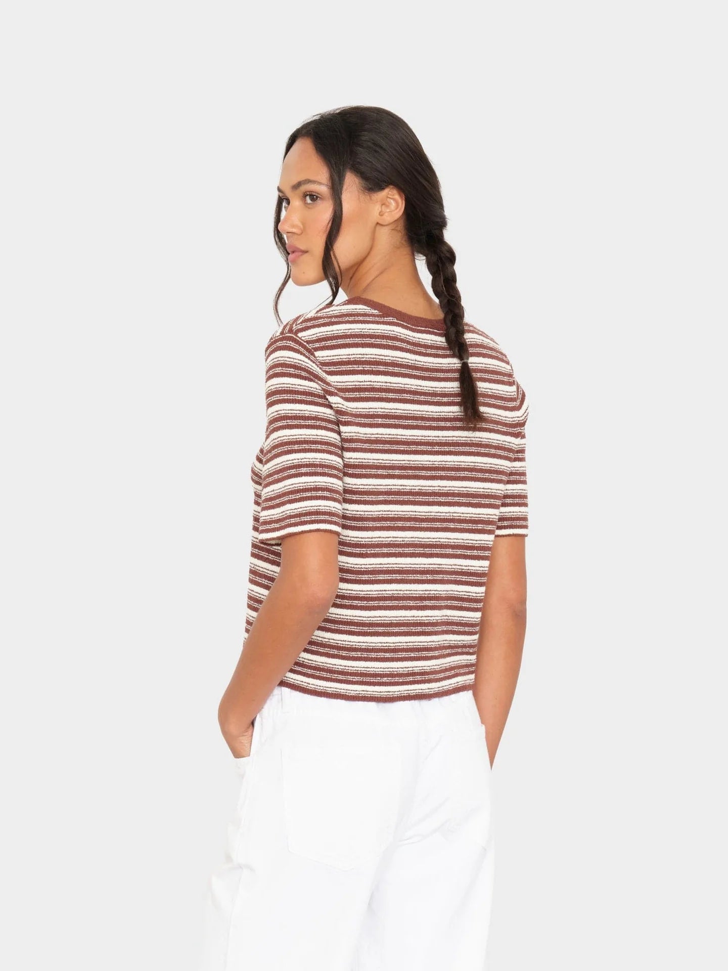 A woman with a single braid stands against a plain light background, hands in pockets, looking over her shoulder. She wears the Xirena Kelsey Top—a brown and white striped, short-sleeve crew neck rib knit top—with white pants.