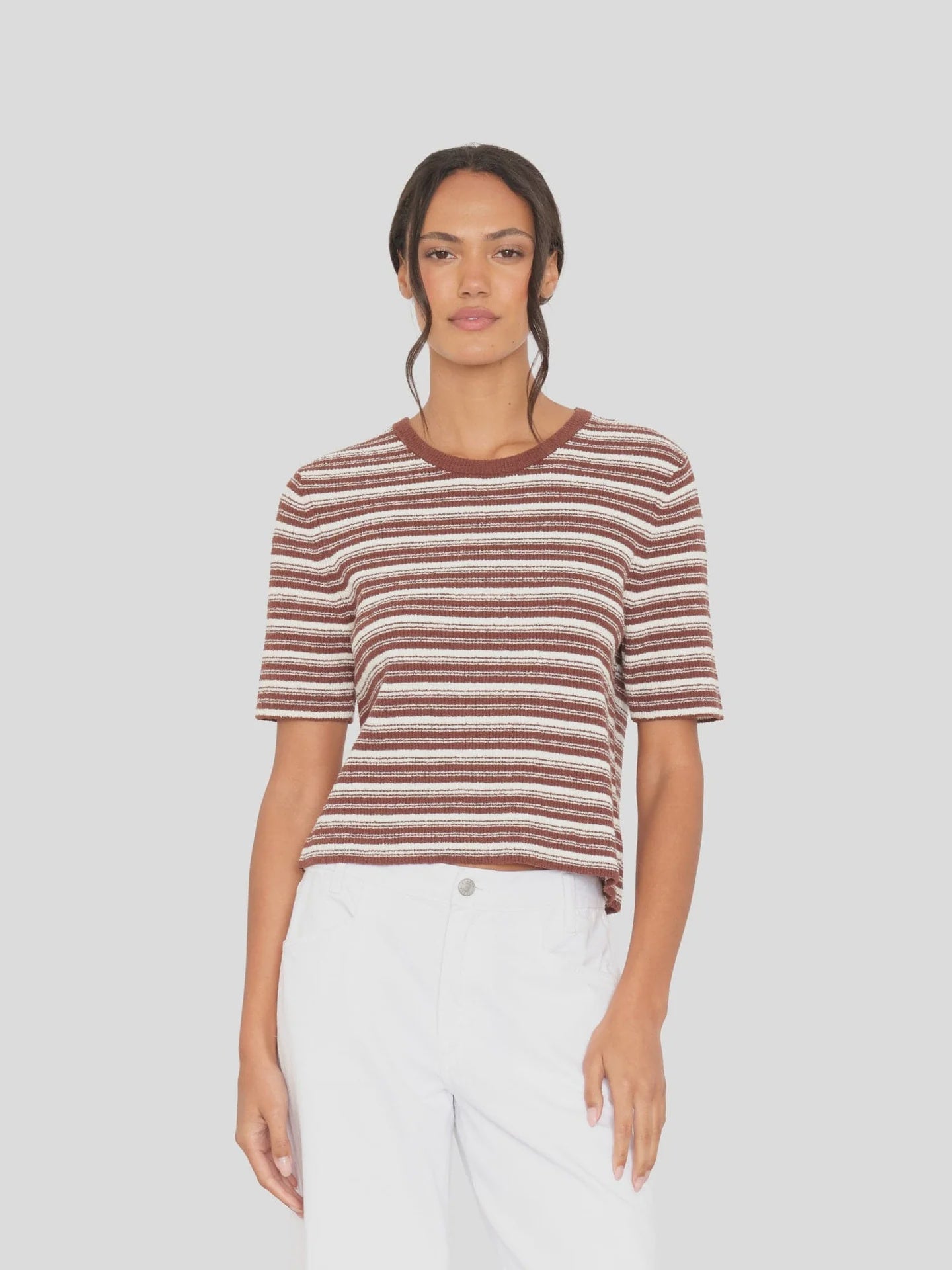 A person wears the Xirena Kelsey Top, a brown and white striped rib knit short-sleeve crew neck, paired with white pants, standing against a plain light grey background and looking into the camera with a neutral expression.
