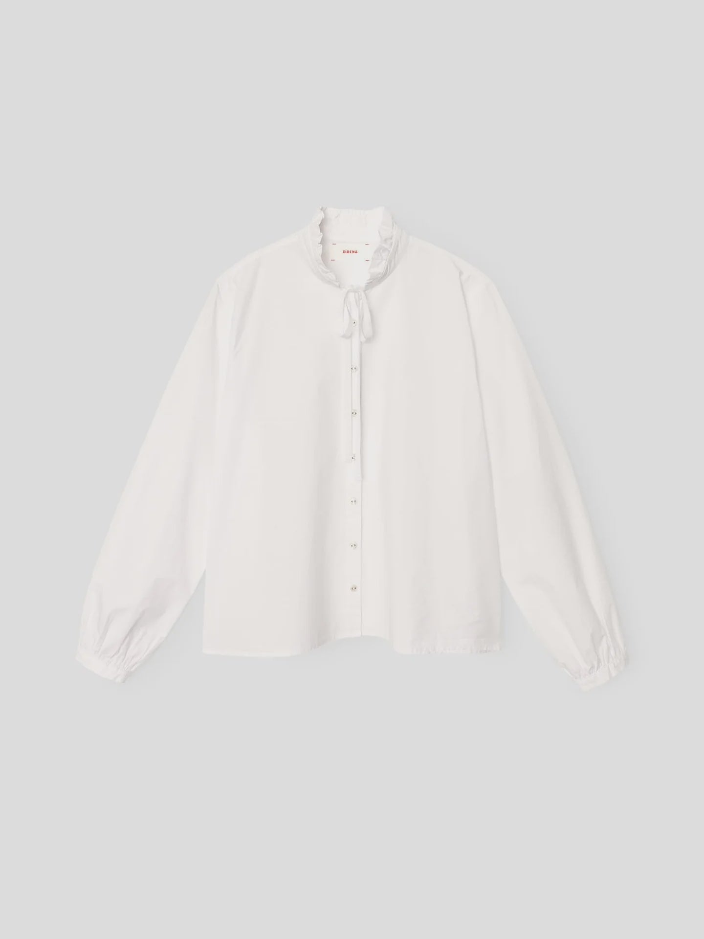 White blouse with ruffled neck, front buttons, and bow by Xirena; Tatiana Shirt in poplin, shown on plain background.