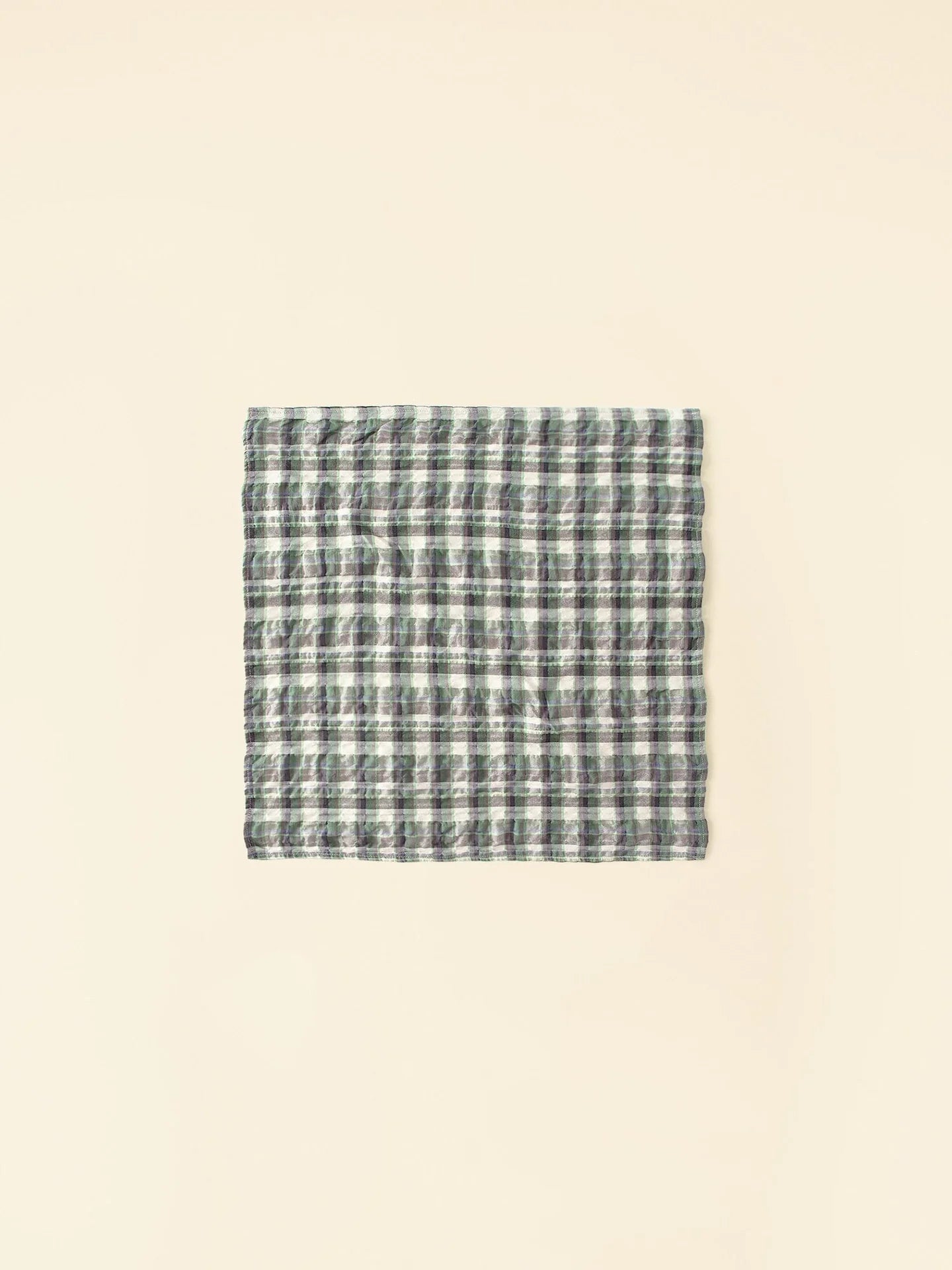 A folded Bandana by Xirena, featuring a checkered pattern with shades of green, gray, and white, displayed against a simple light beige background.