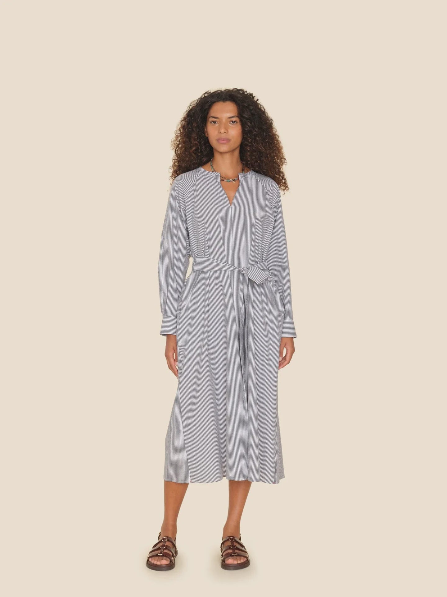 A woman with curly hair stands against a beige background, wearing the Sylvie Dress by Xirena. The dress is a V-neck midi style with cotton pencil stripes and a self-tie belt at the waist. She completes her look with brown sandals.